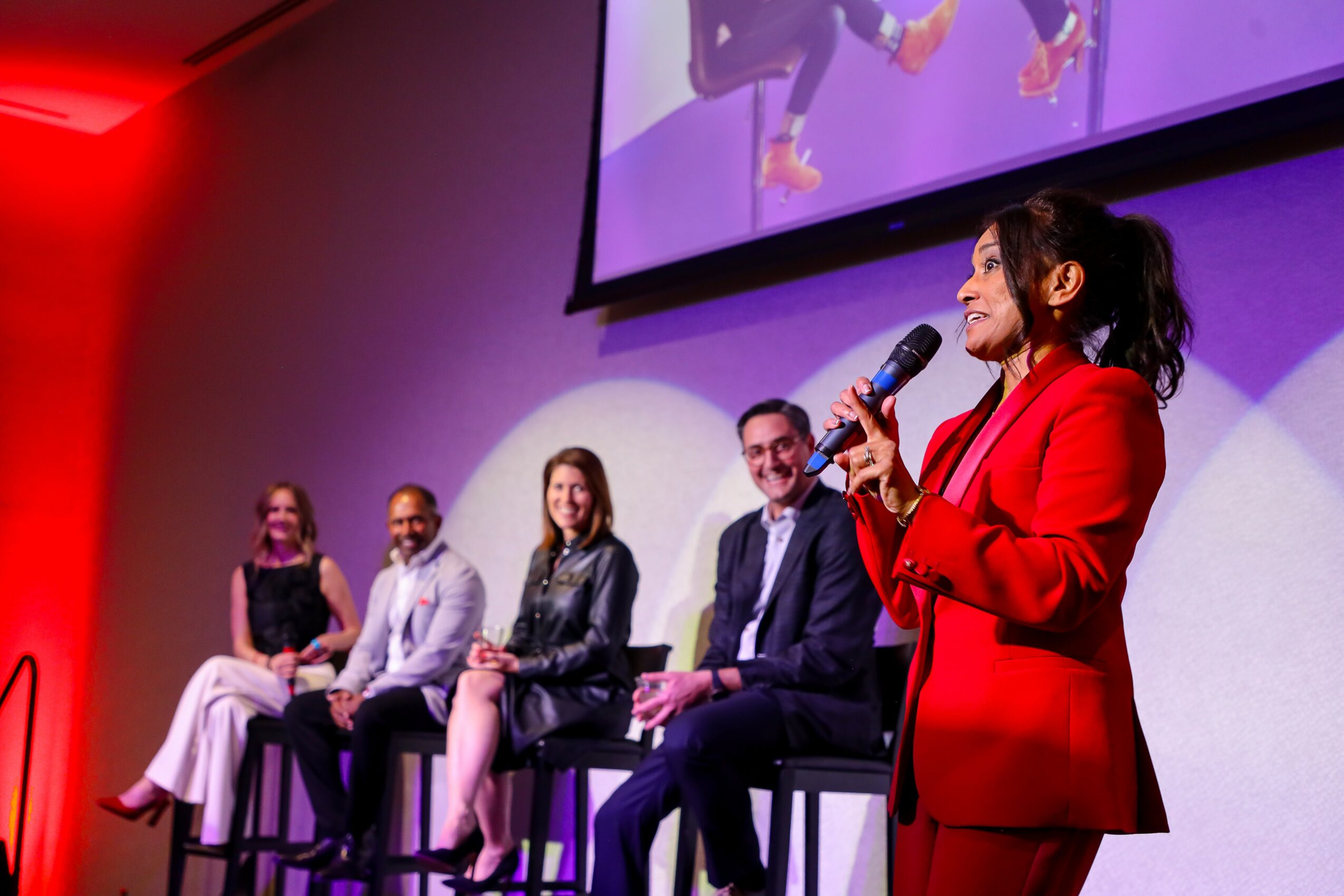 Woman in red blazer speaking into microphone on stage with four seated people behind her, large screen above showing her image.