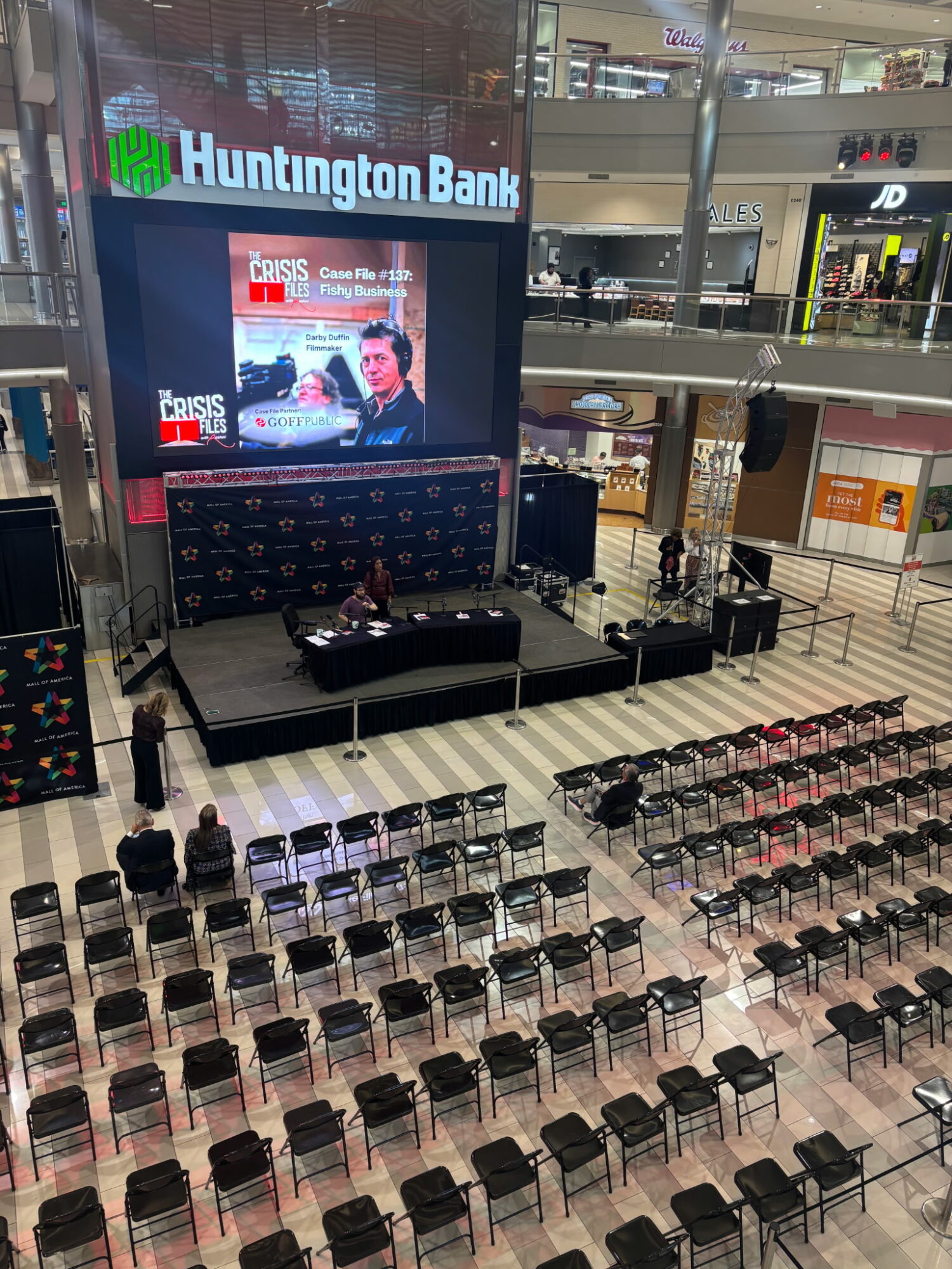 Empty chairs arranged in rows in a shopping mall, stage with a large screen and Huntington Bank sign above.