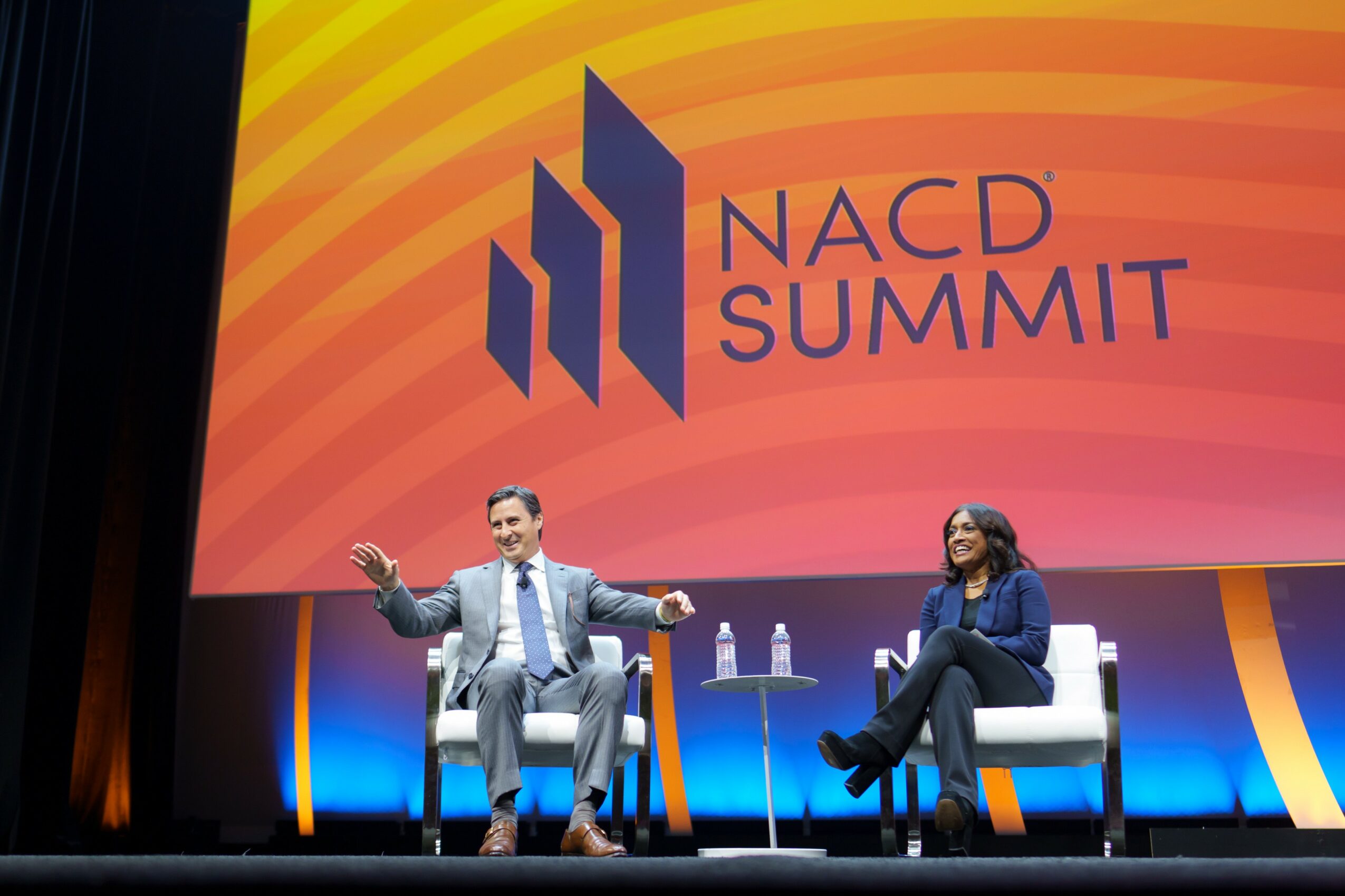 Two people seated on stage in front of a large NACD Summit screen, one speaking and gesturing, the other listening.