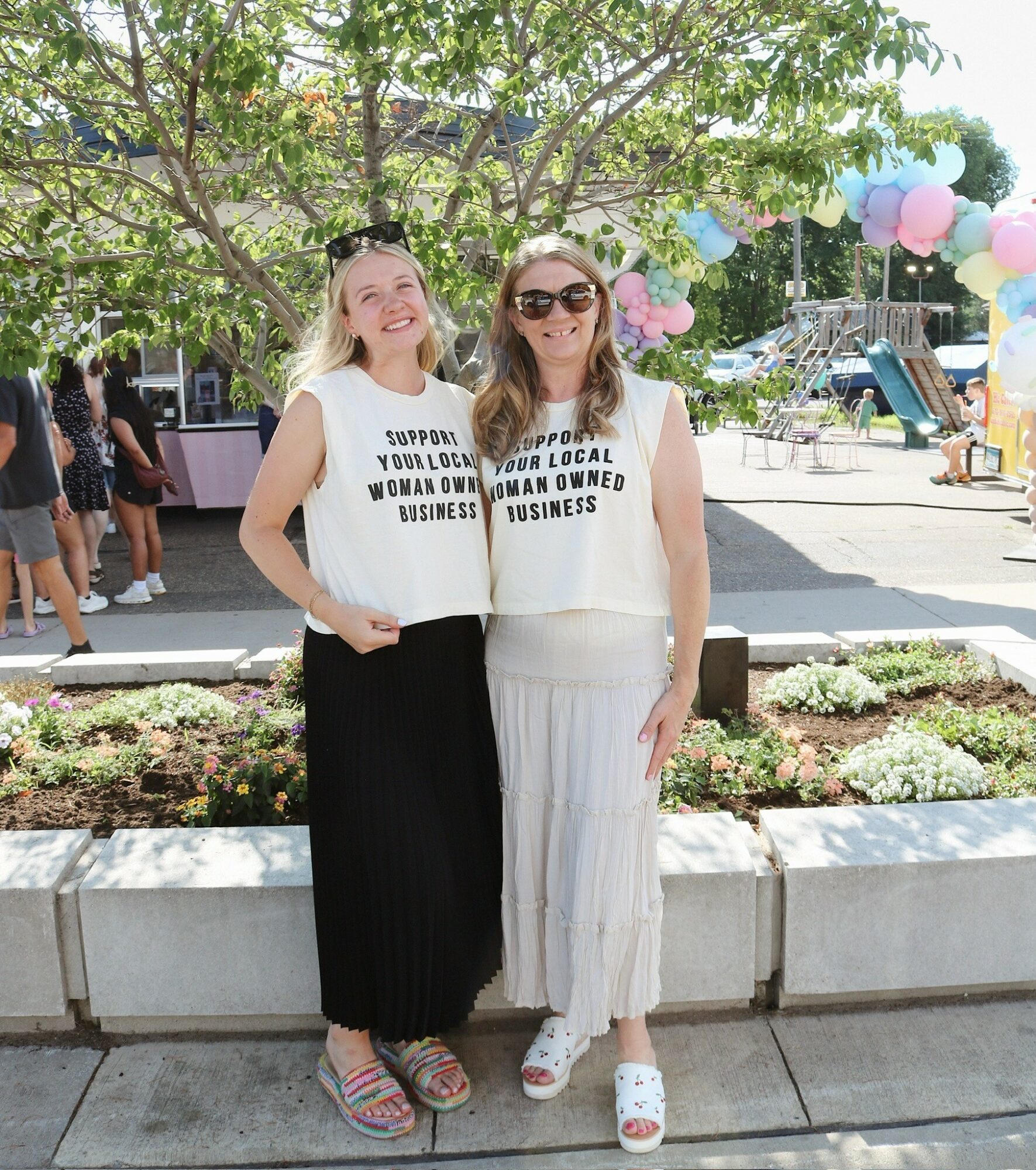 Two women standing outdoors in front of a tree, smiling, wearing casual clothing, with balloons and people in the background.
