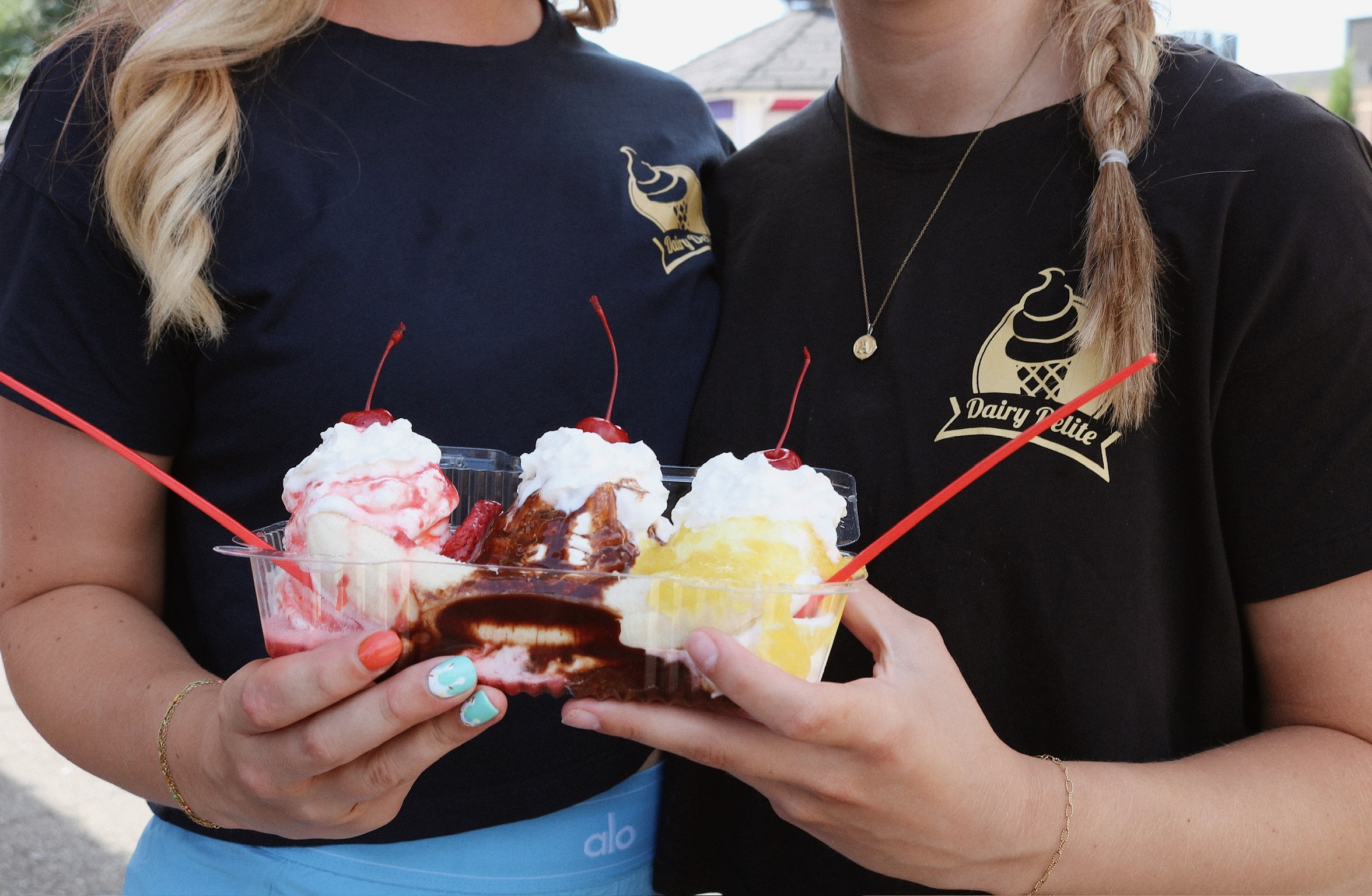 Two people holding a tray of ice cream sundaes with whipped cream, berries, and toppings, wearing black shirts with a logo.