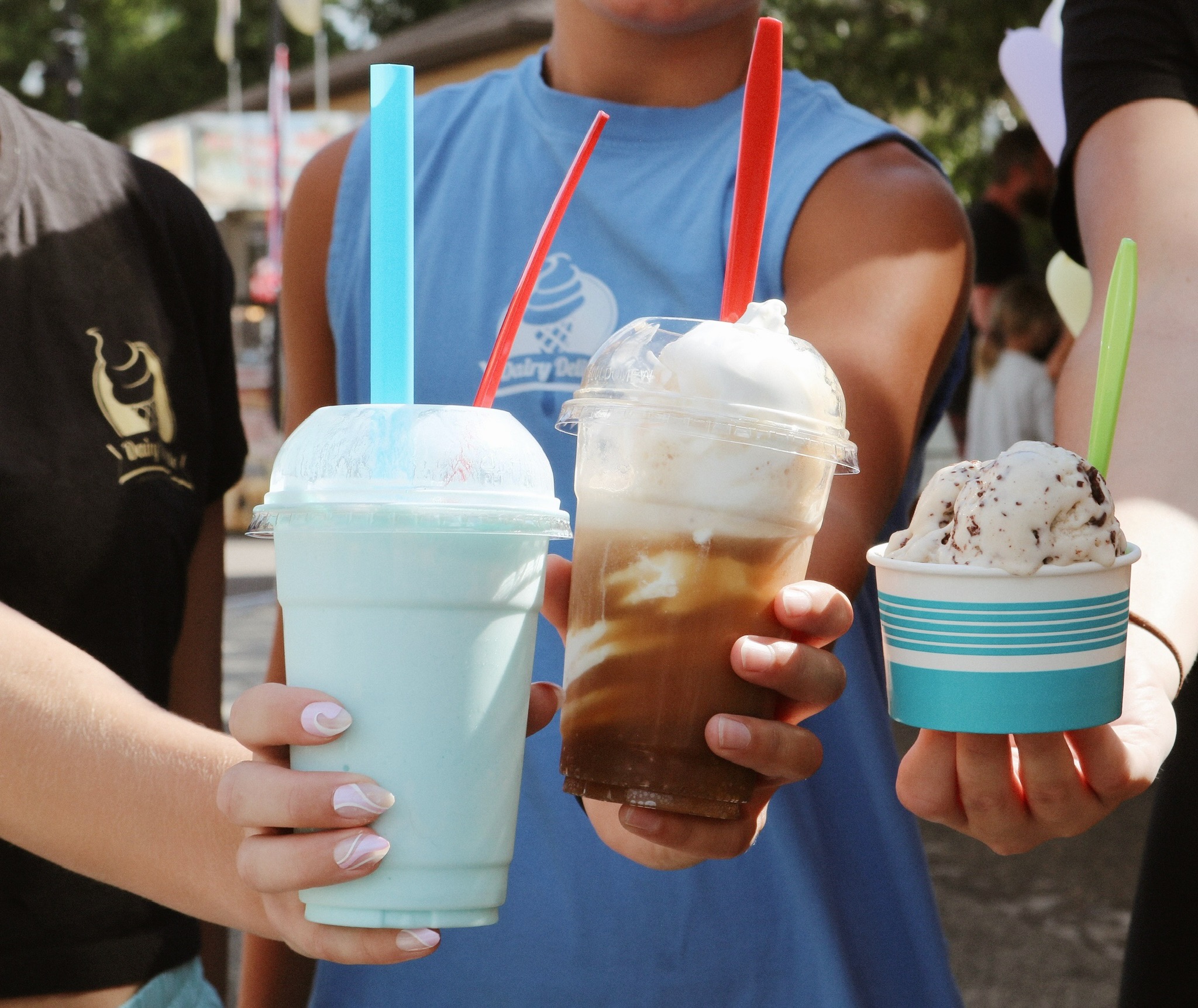 Three people holding colorful drinks with straws, outdoors, with trees and people in the background.
