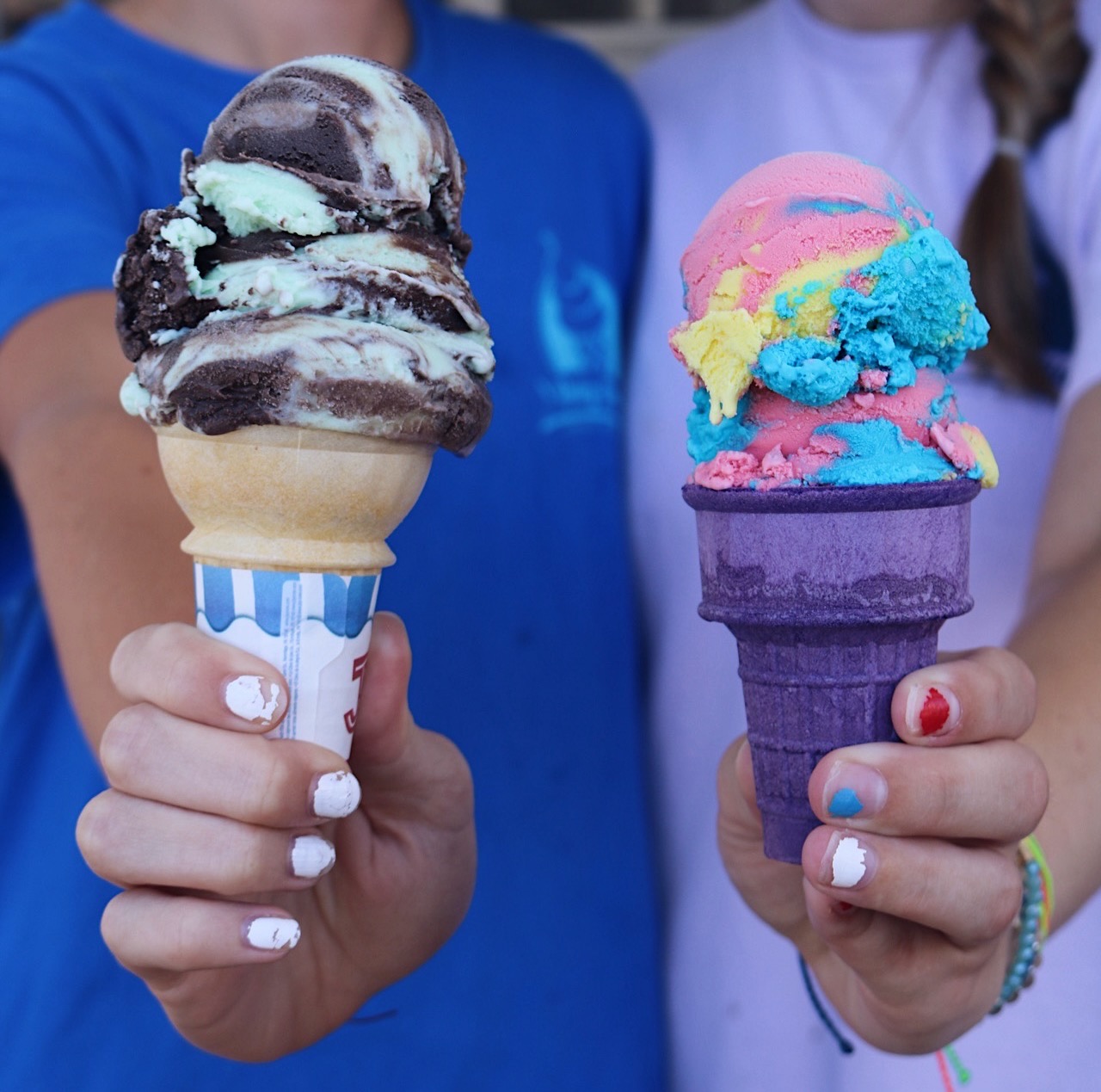 Two people hold ice cream cones with multiple scoops, one with chocolate and vanilla, the other with colorful rainbow flavors.
