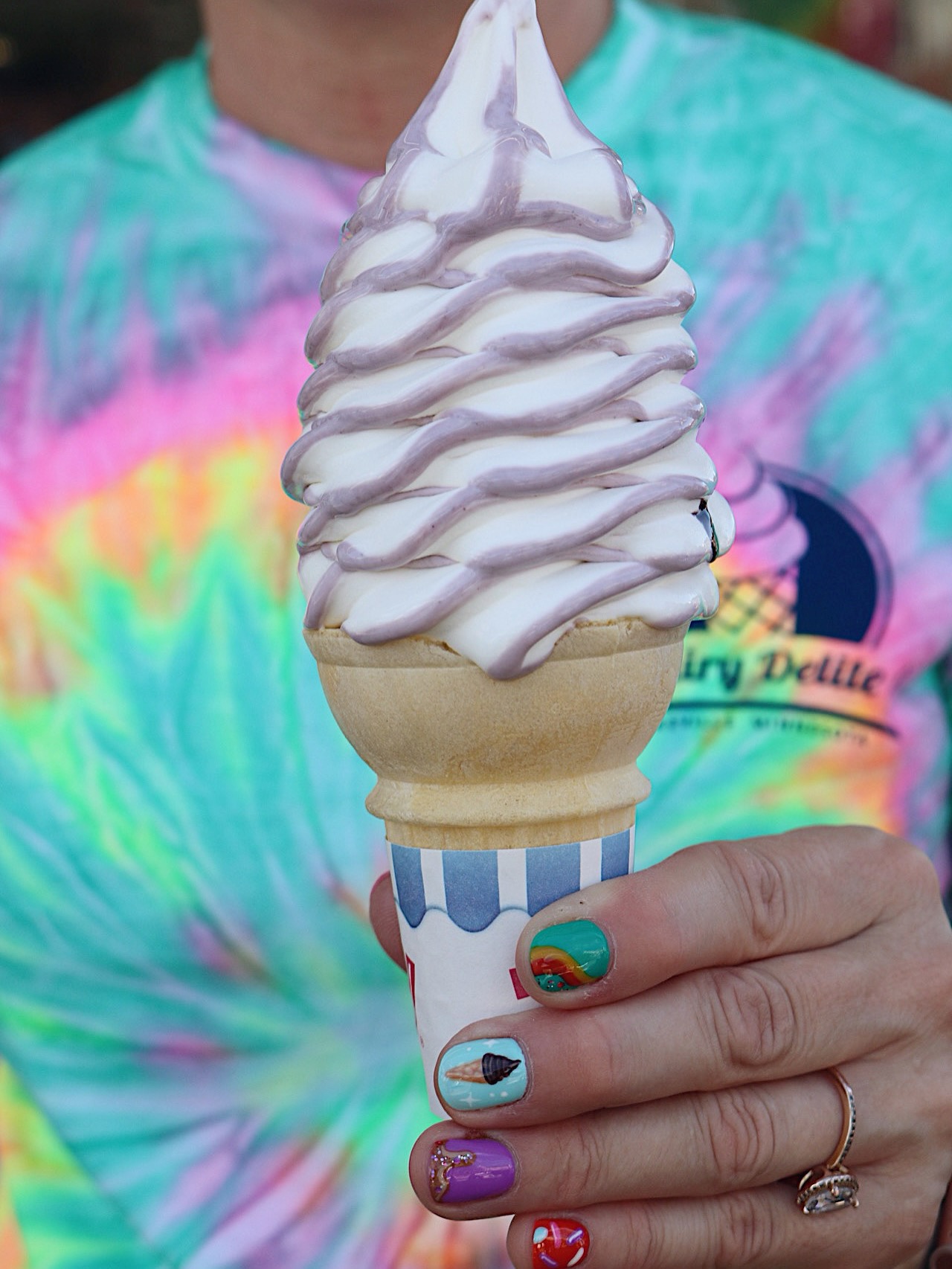 Person holding a soft-serve ice cream cone with a colorful tie-dye shirt visible in the background.
