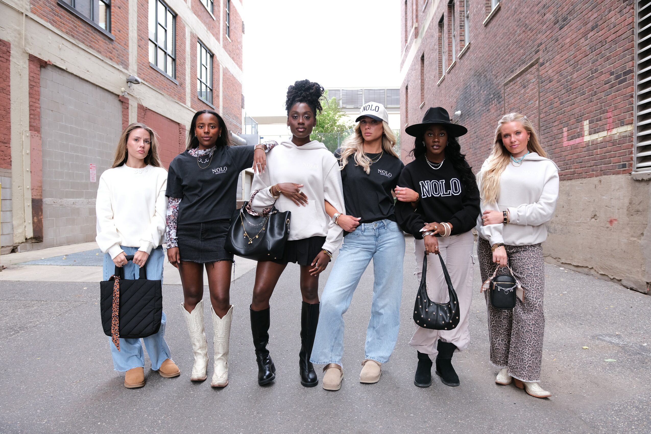 Six women standing in an alleyway between brick buildings, dressed in casual and stylish clothing, holding bags.
