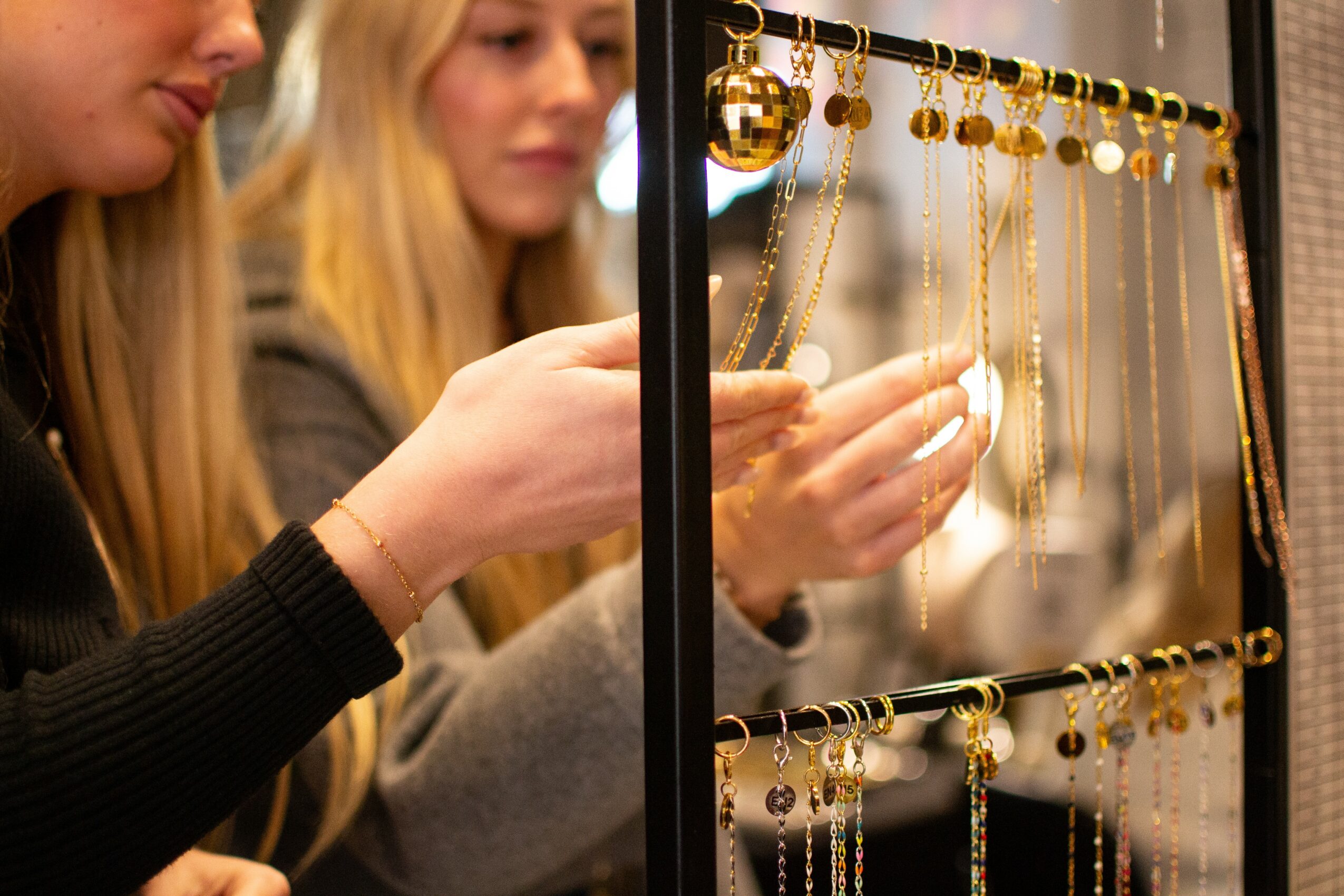 Two women look at gold jewelry displayed on a rack, with one woman touching a necklace. Focus on jewelry display.