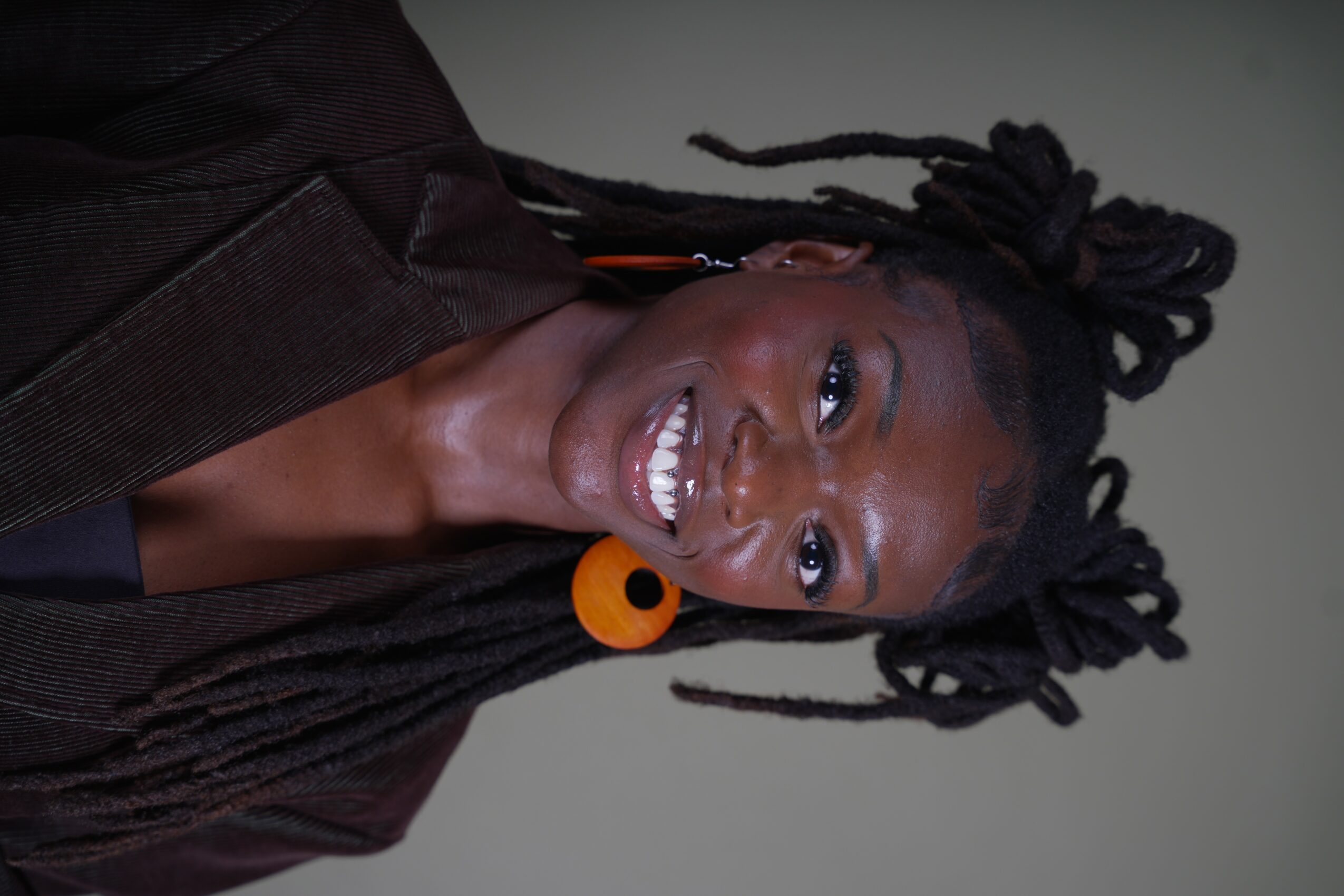 Woman with dreadlocks and orange earrings smiling, wearing a dark jacket, against a plain background.