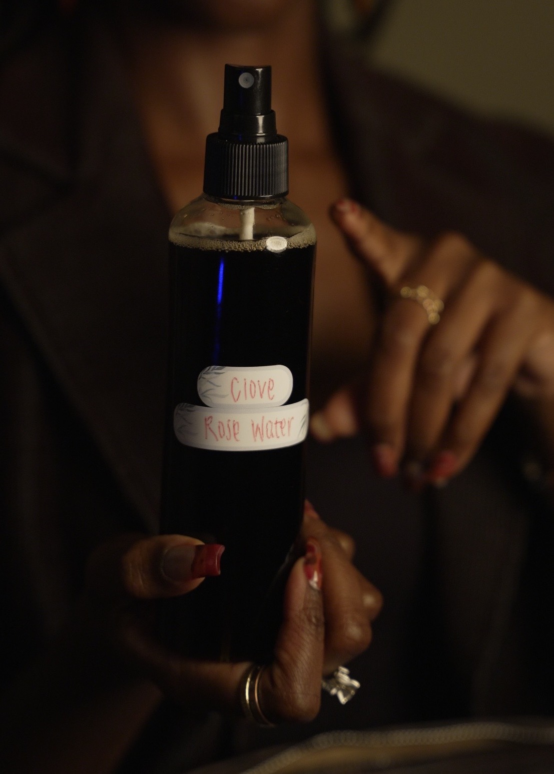 Person holding a black spray bottle labeled 'Clove' and 'Rose Water' with a hand showing a ring and painted nails.