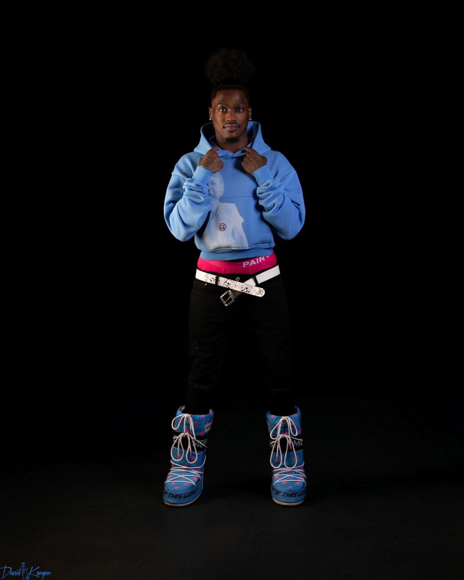 Young person standing with hands near face, wearing a blue hoodie, black pants, and colorful sneakers, against a black background.
