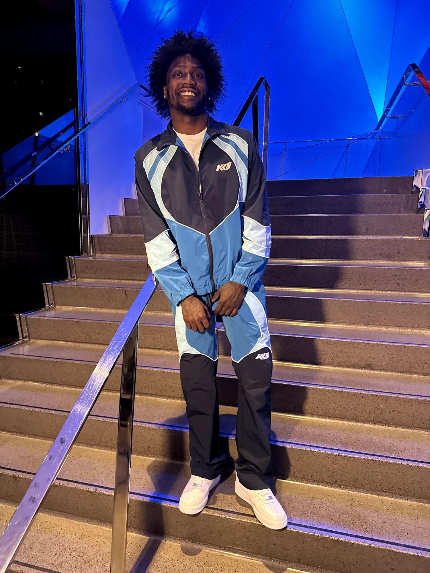 Person with curly hair in a blue, black, and white jacket standing on stairs with a blue-lit background.