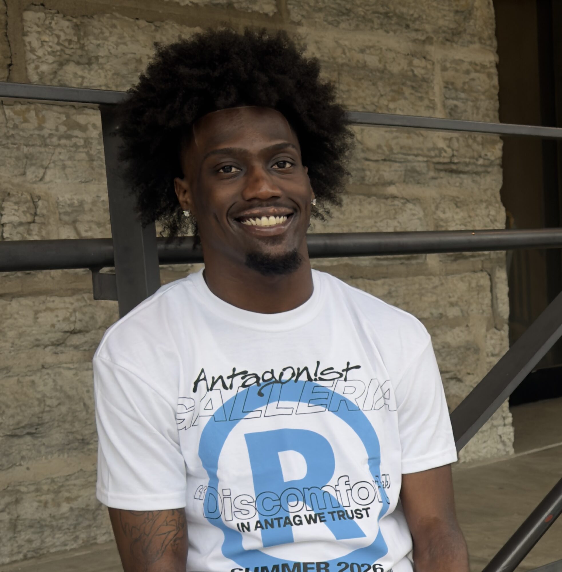 Young man with curly hair smiling, wearing a white T-shirt with blue and black text, standing outdoors against a stone wall.