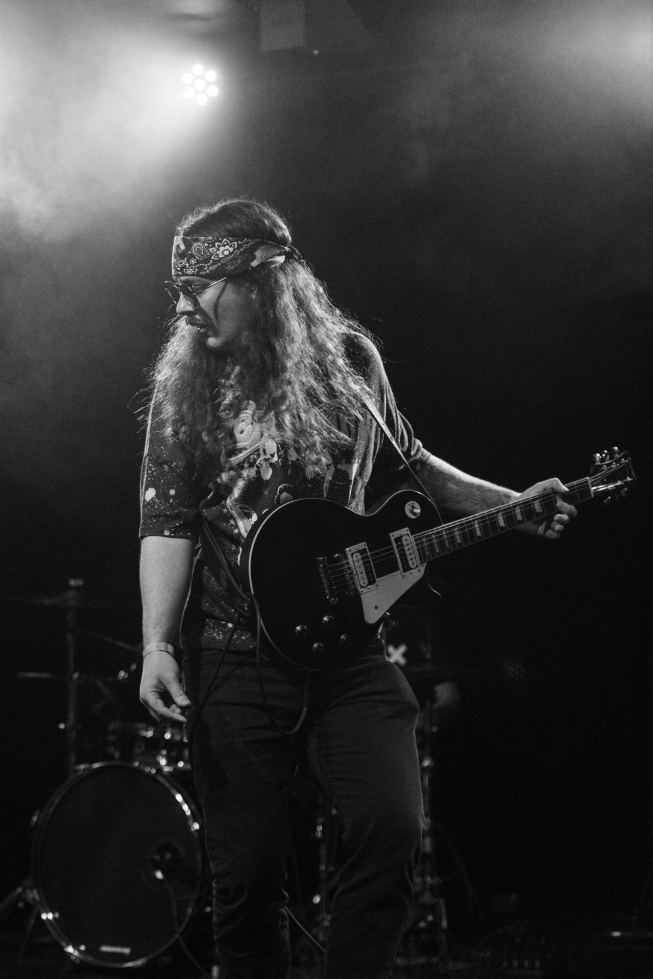 Person with long hair and headband holding an electric guitar on stage, with a drum set in the background, in black and white.