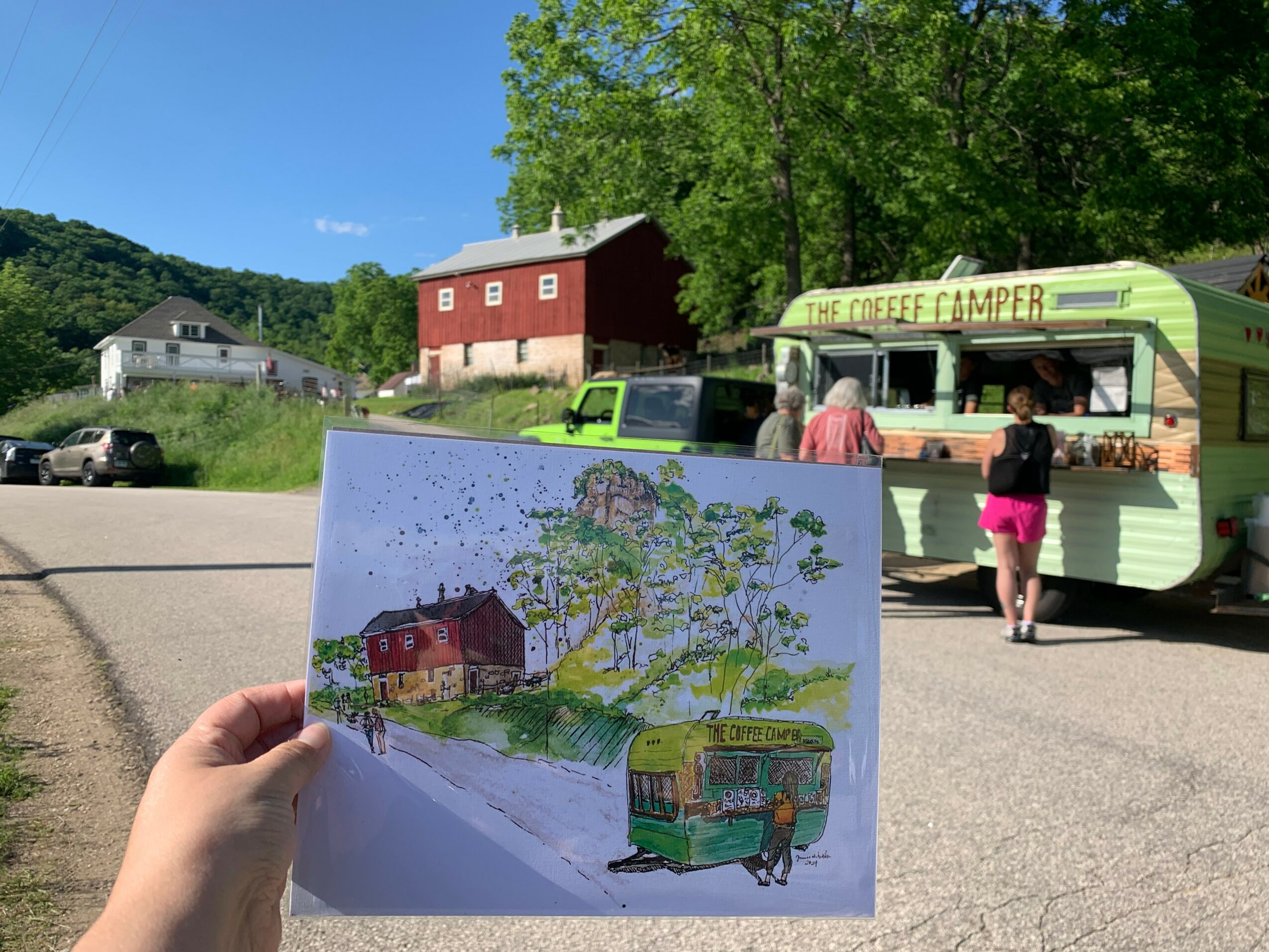 Person holding a colorful drawing of a red barn, trees, and a bus, with a food truck and people in the background.