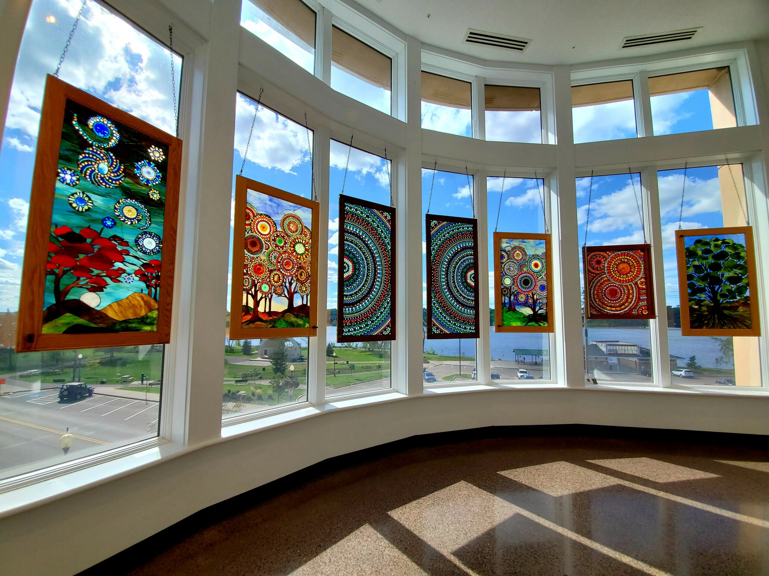 Gallery with colorful paintings on windows, sky and clouds visible outside, curved wall, wooden floor, bright natural light.