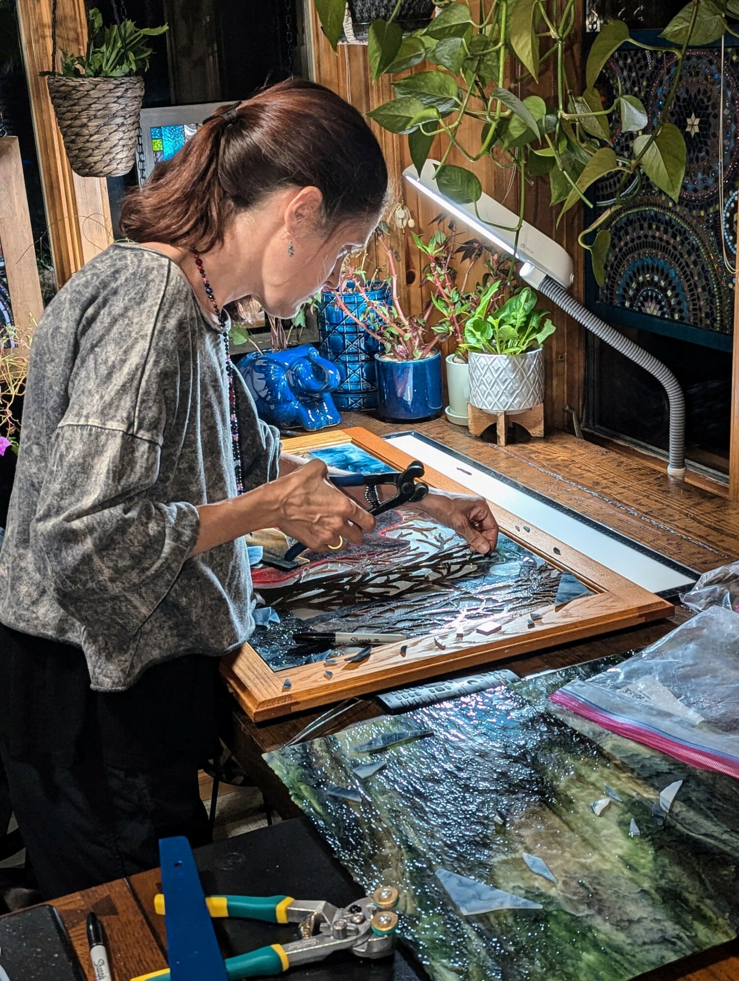 Woman working on a framed artwork at a wooden table with plants and art supplies nearby.