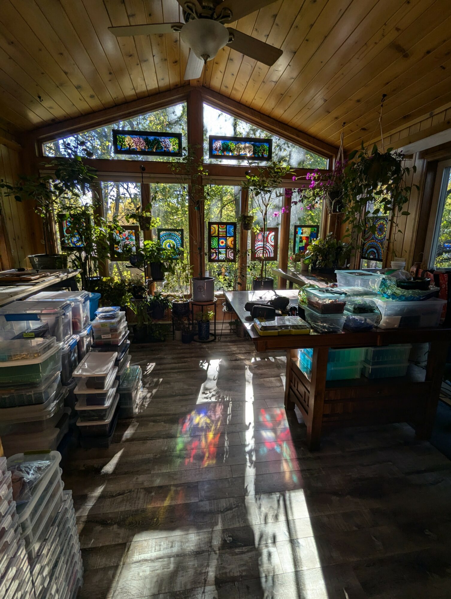 Interior of a room with wooden walls and ceiling, large stained glass windows, plants, and tables with storage containers.