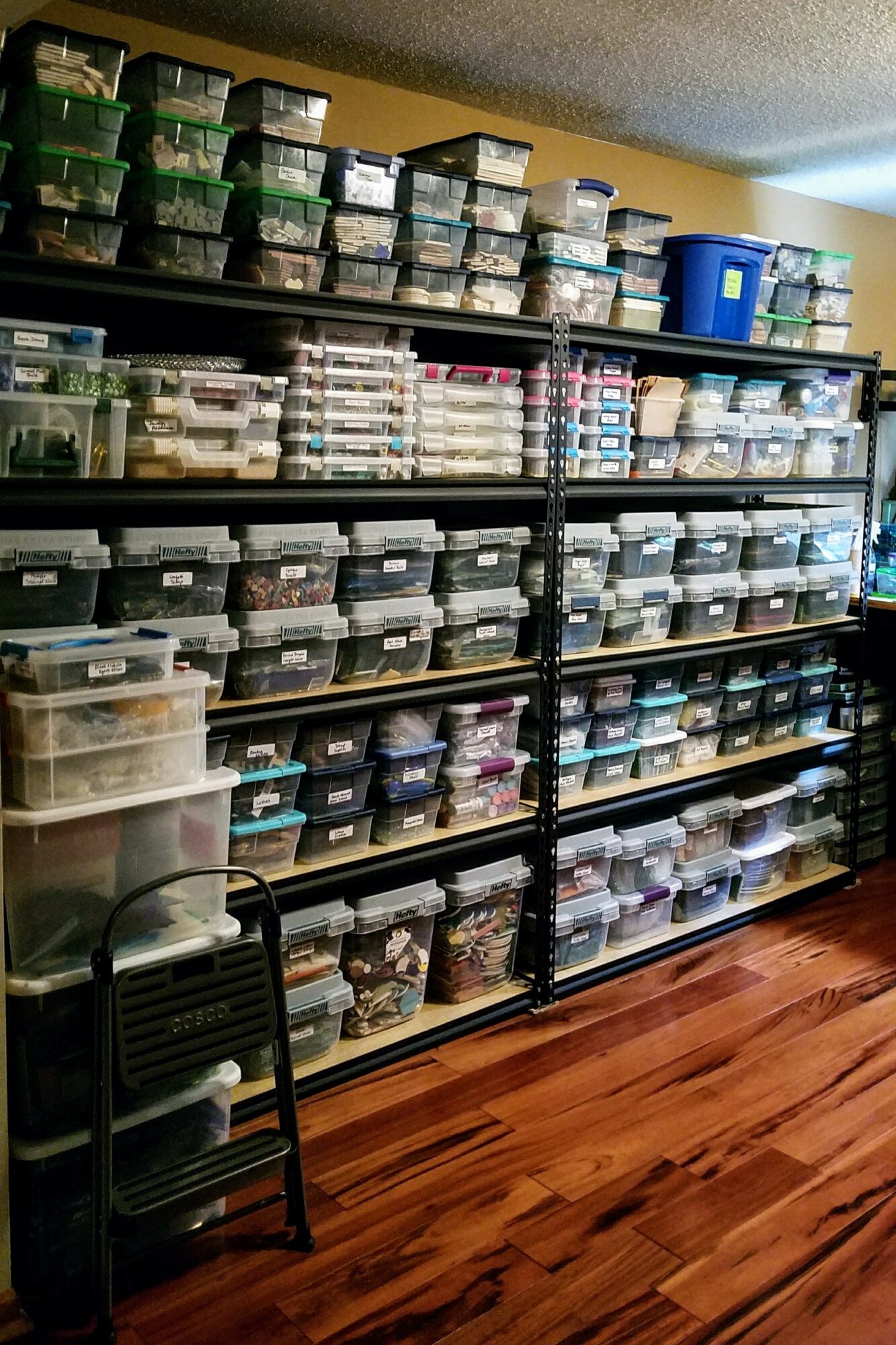 Black metal shelving unit filled with plastic containers and boxes, with a small black chair in front, on a wooden floor.