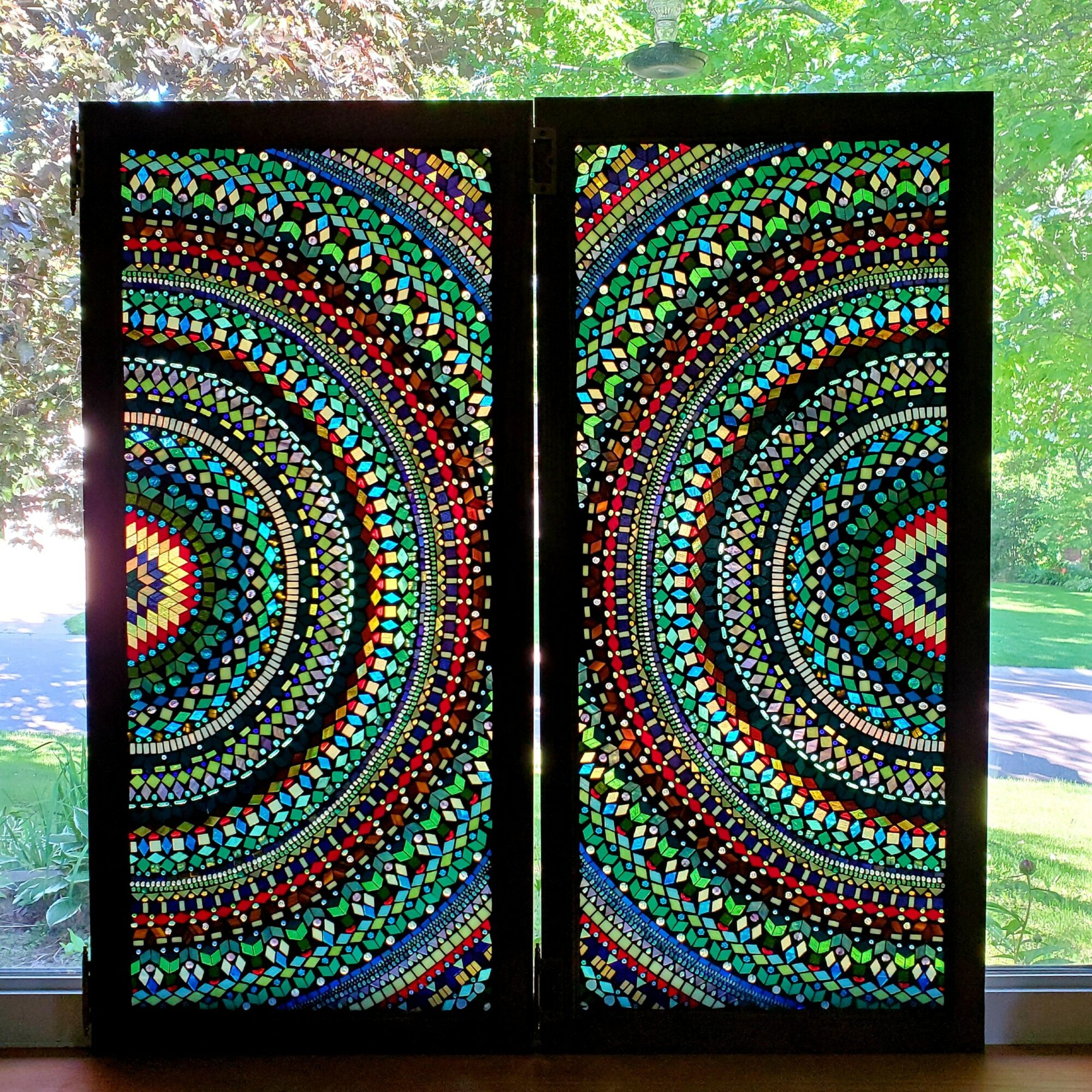 Colorful mosaic artwork with concentric circles on two panels, set against a window with trees outside.