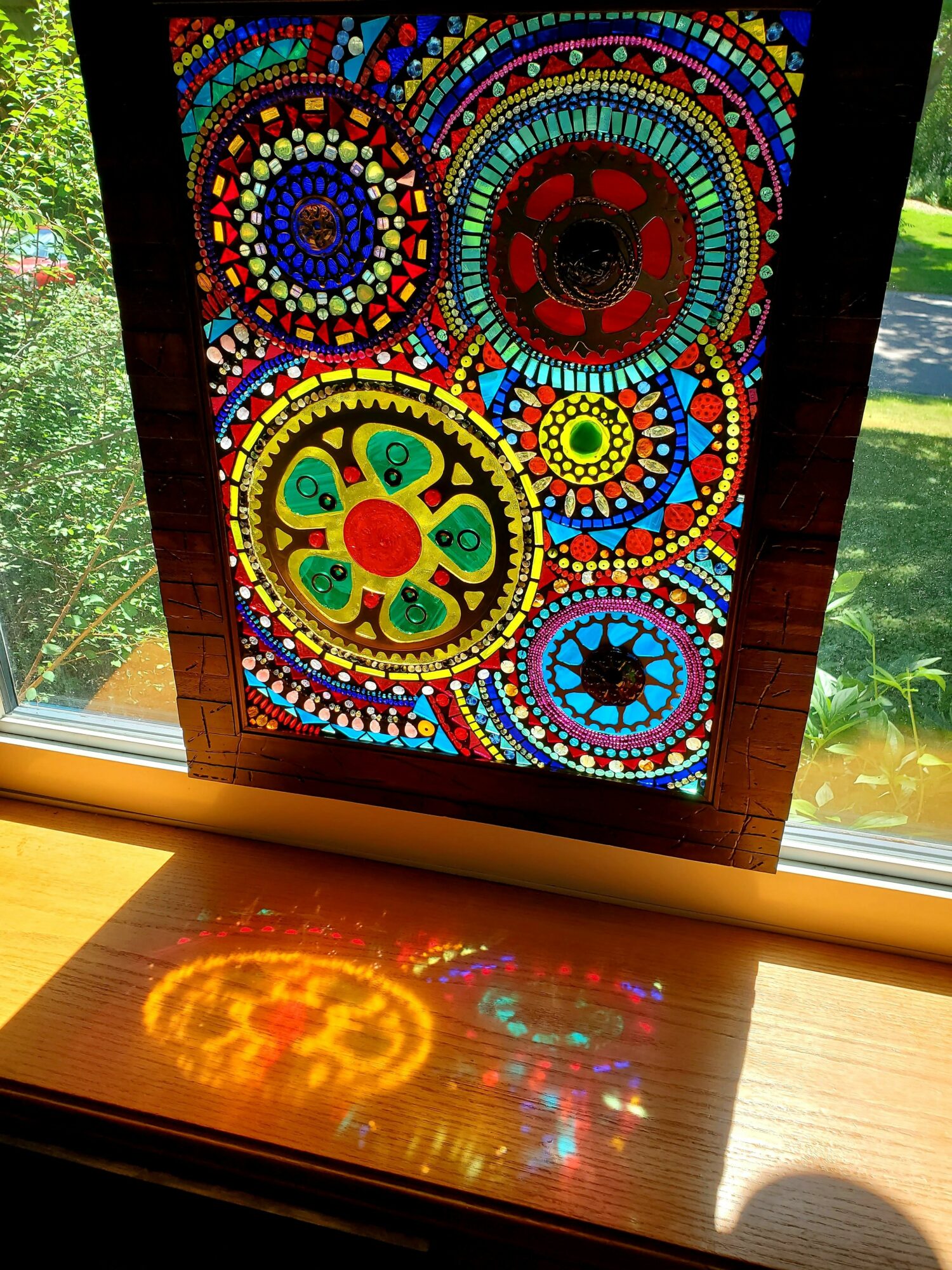 Colorful mosaic stained glass window with circular patterns, sunlight casting reflections on a wooden surface, outdoors visible through window.