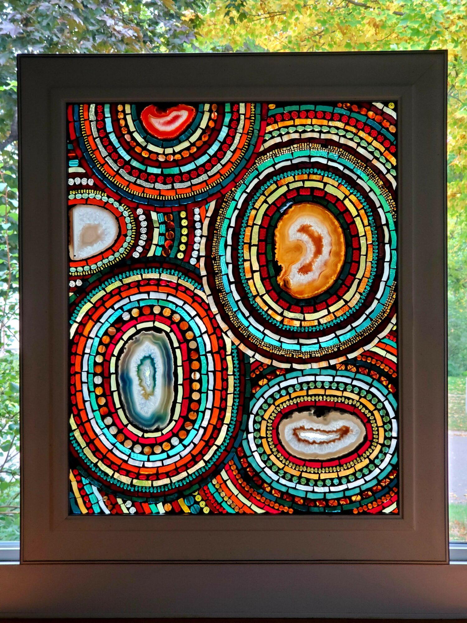 Colorful mosaic pattern with concentric circles and various shapes, framed in black, outdoors with trees and leaves visible.