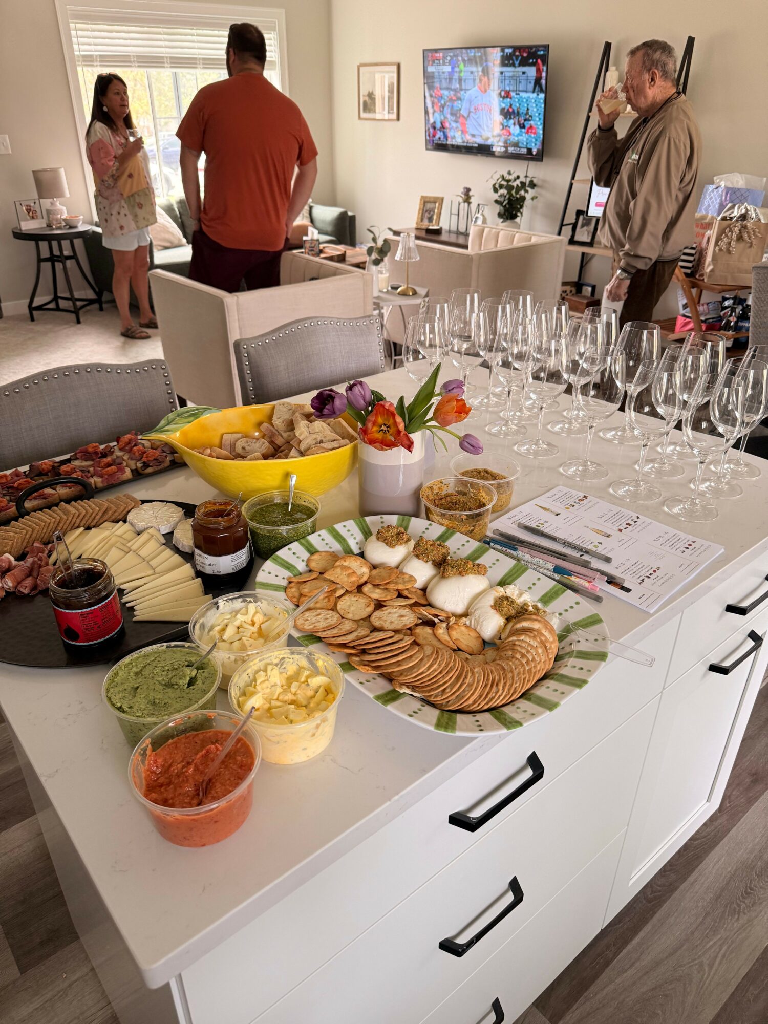 Table with snacks, drinks, and a floral centerpiece in a room with people and a TV screen.