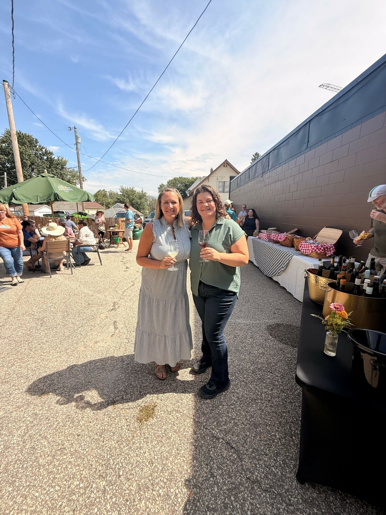 Two women standing outdoors near a market stall, with people and a building in the background.