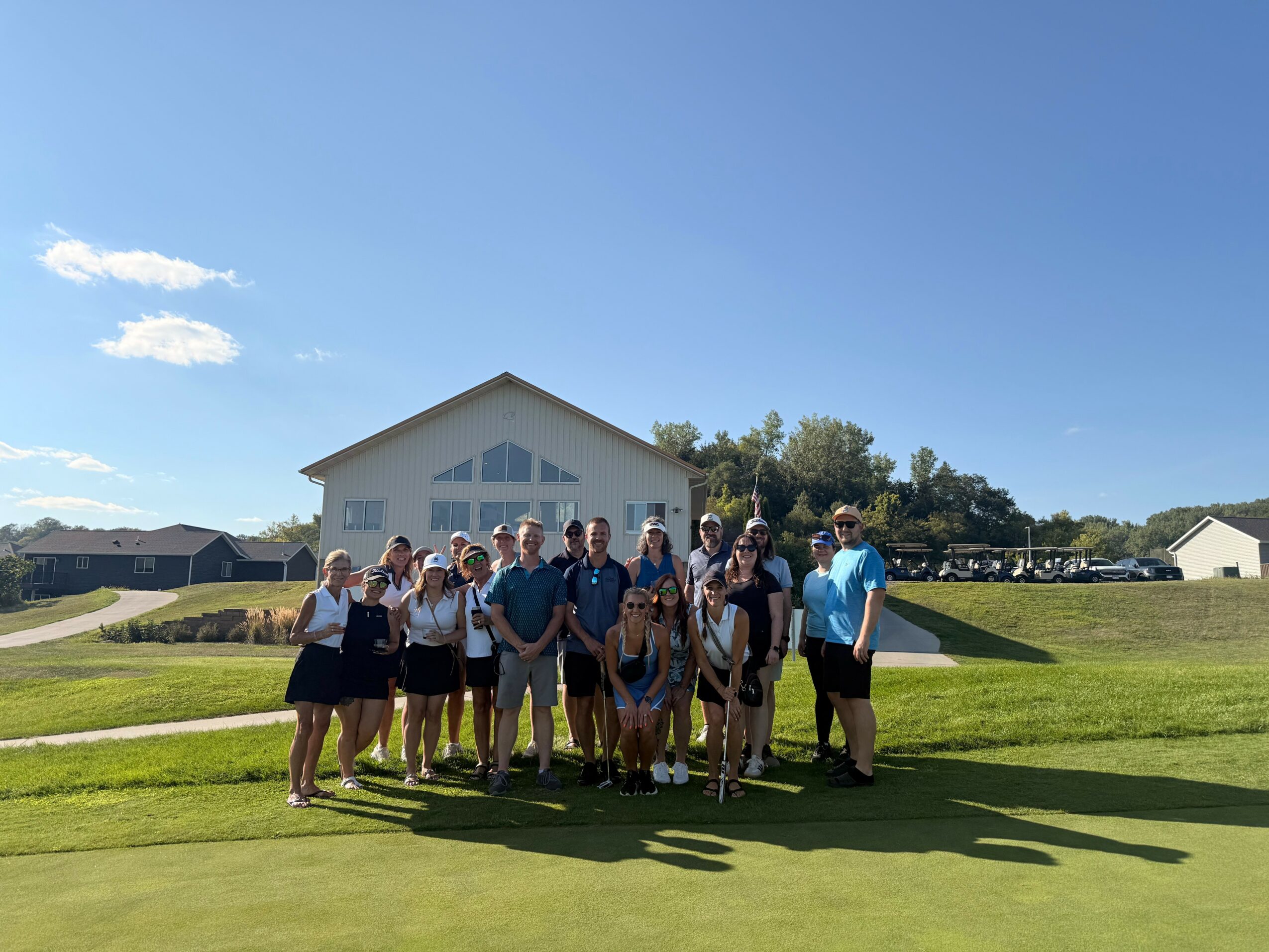 Group of people standing outdoors on grass in front of a white building with a blue sky background.