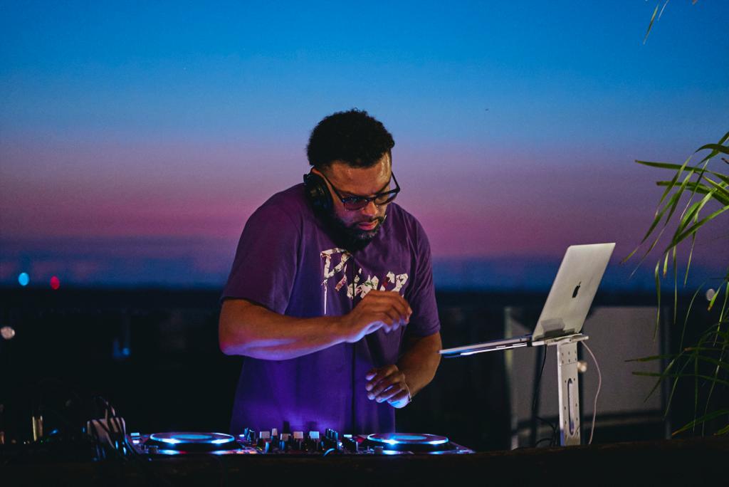 DJ wearing glasses and a purple shirt, operating equipment outdoors during sunset with a laptop and plants nearby.