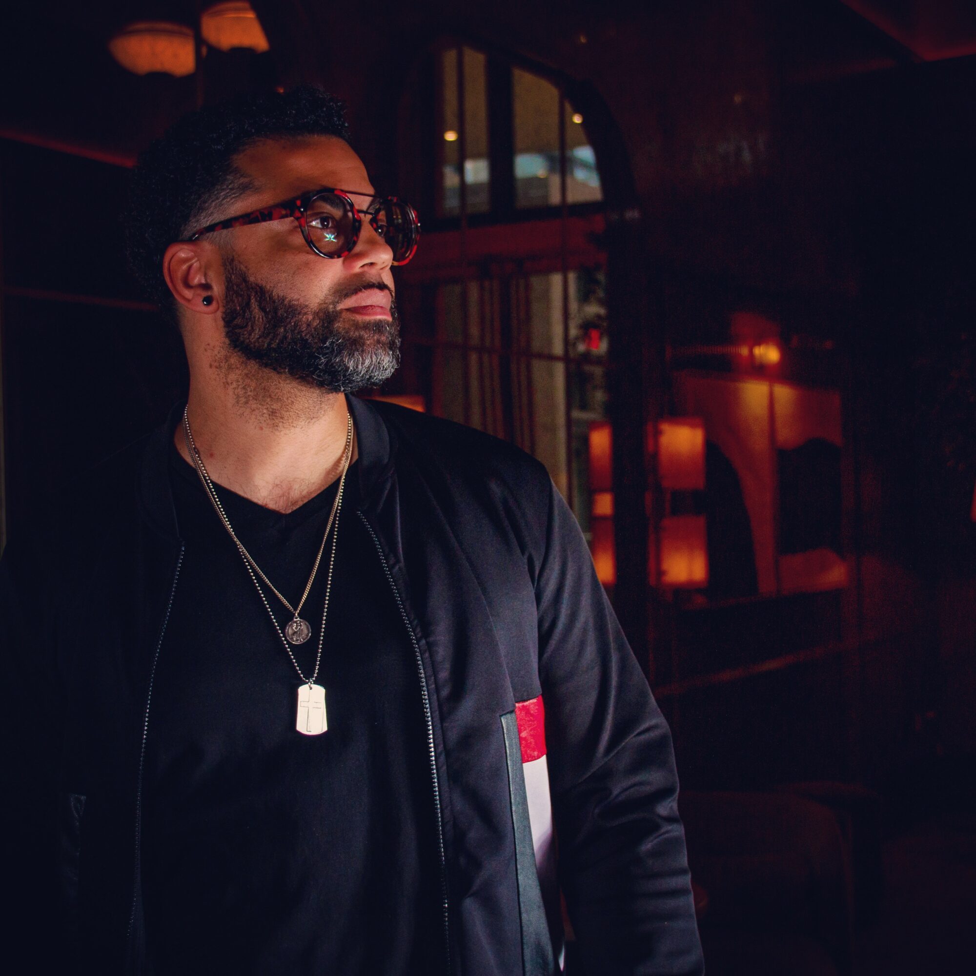 Man with glasses and beard wearing a black jacket in a dimly lit setting.