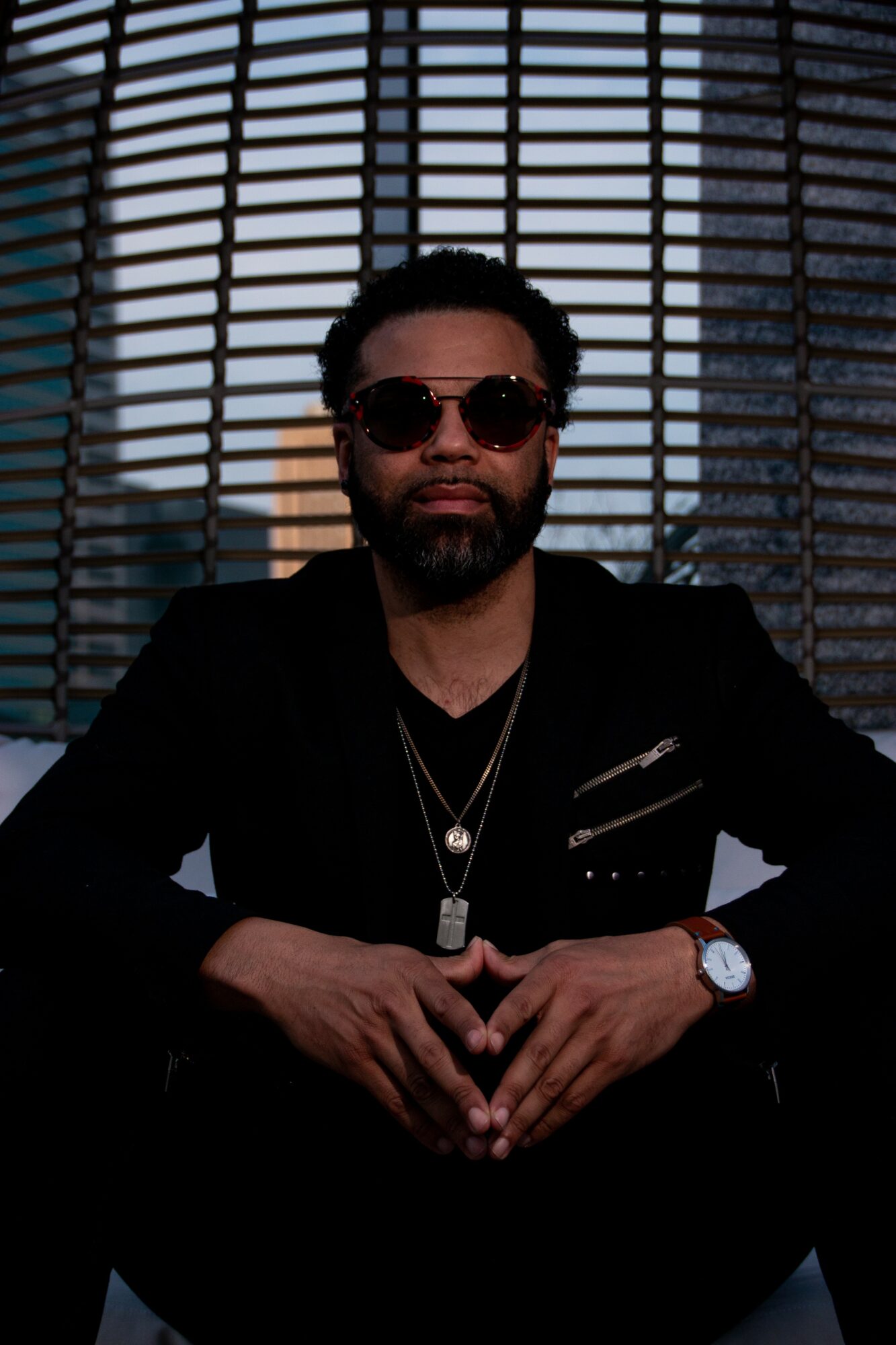 Man with sunglasses and beard sitting in front of a window with blinds, wearing dark clothing and jewelry.
