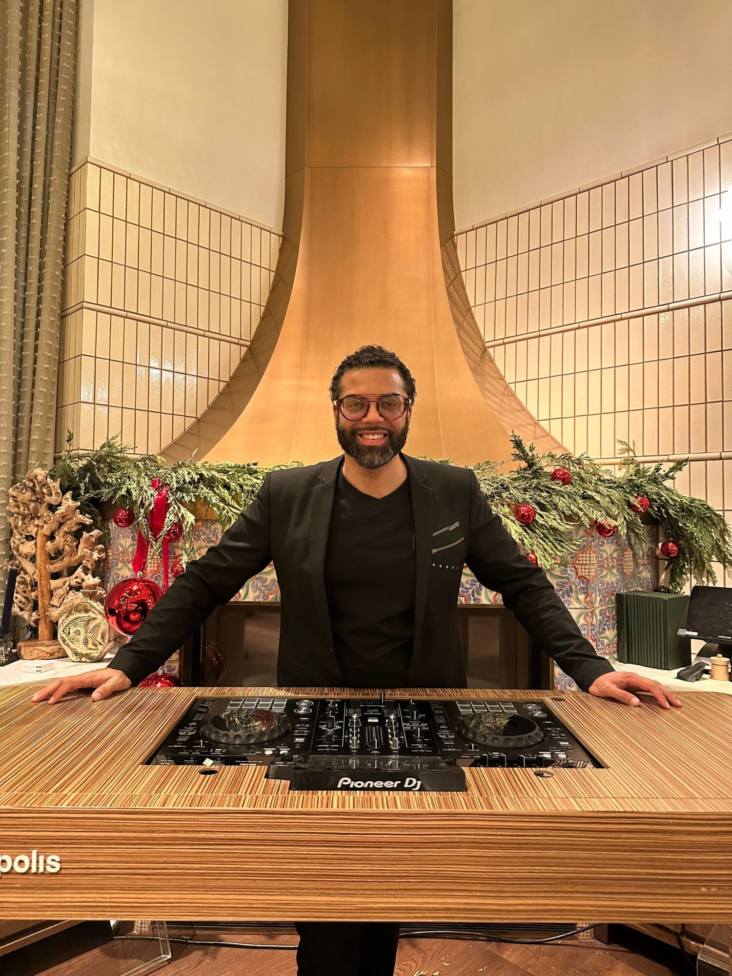 Man with glasses in black suit standing behind a table with a DJ mixer, decorated with Christmas flowers and ornaments.