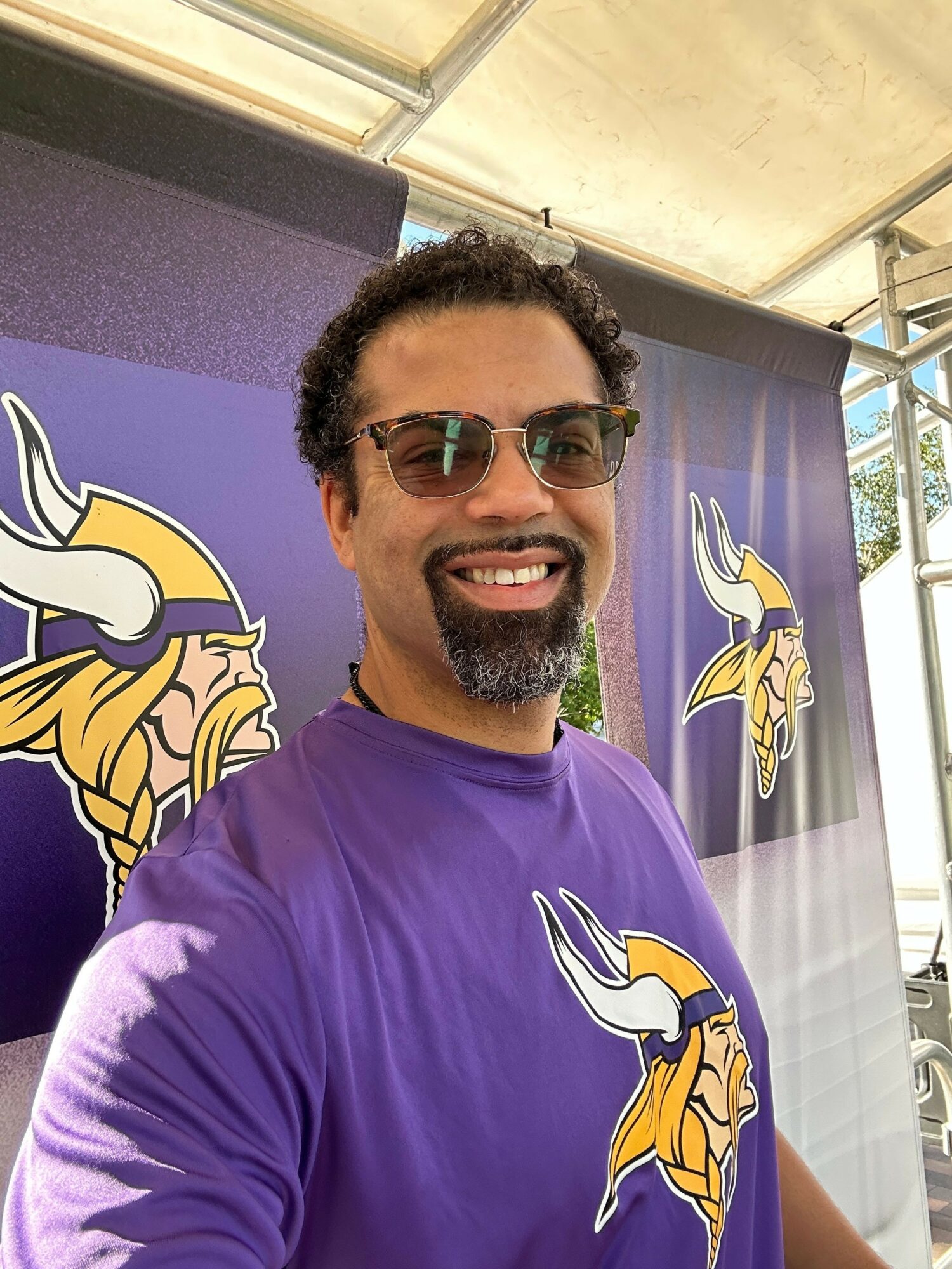 Smiling man with sunglasses and curly hair in a purple shirt, standing in front of a Vikings logo banner.