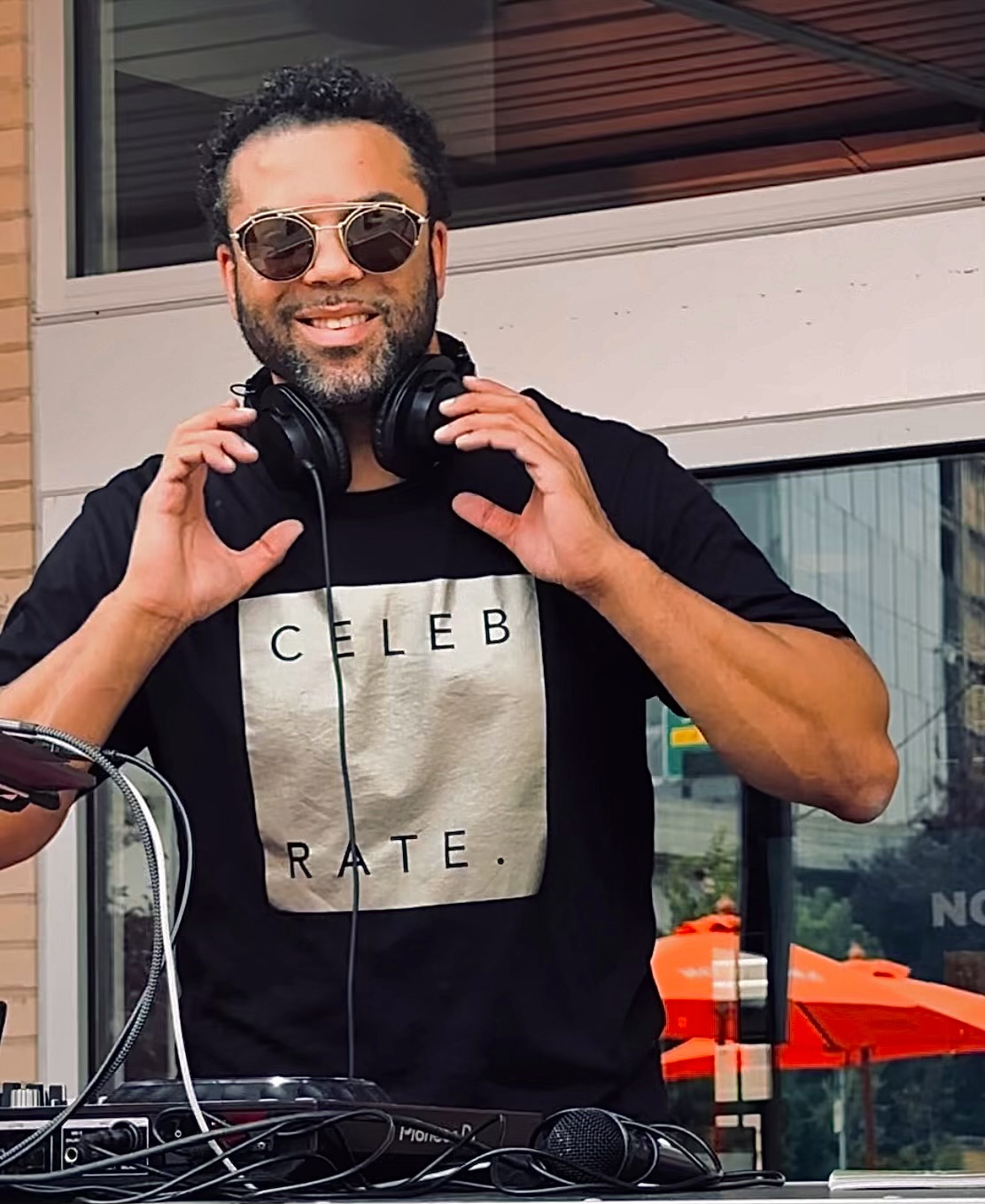 Man with sunglasses and headphones smiling outdoors, wearing a black T-shirt with text, standing near a window.