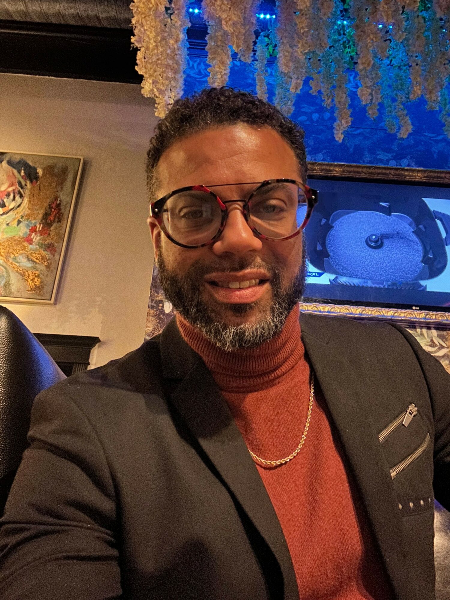 Man with glasses and beard smiling, wearing a black jacket and red shirt, indoors with a painting and window behind him.