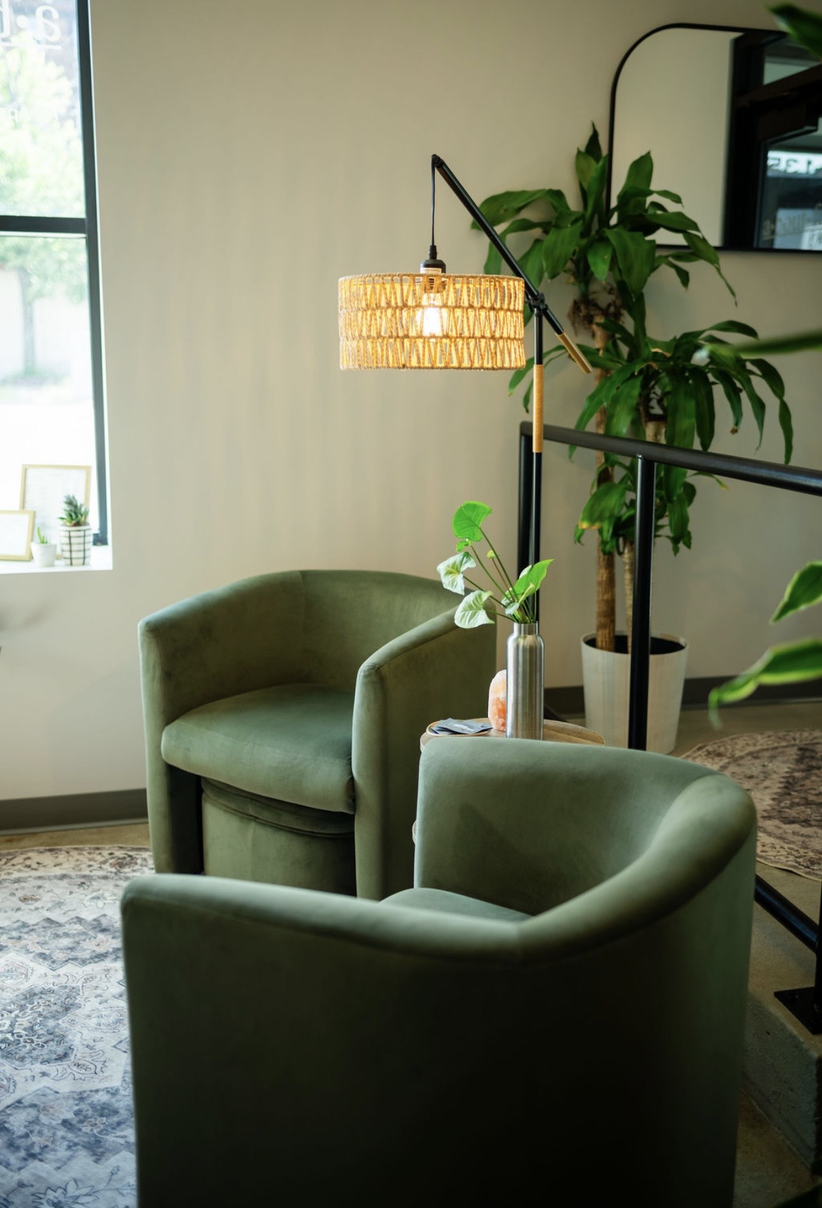 Two green armchairs in a room with a window, a tall plant, and a floor lamp with a woven shade.