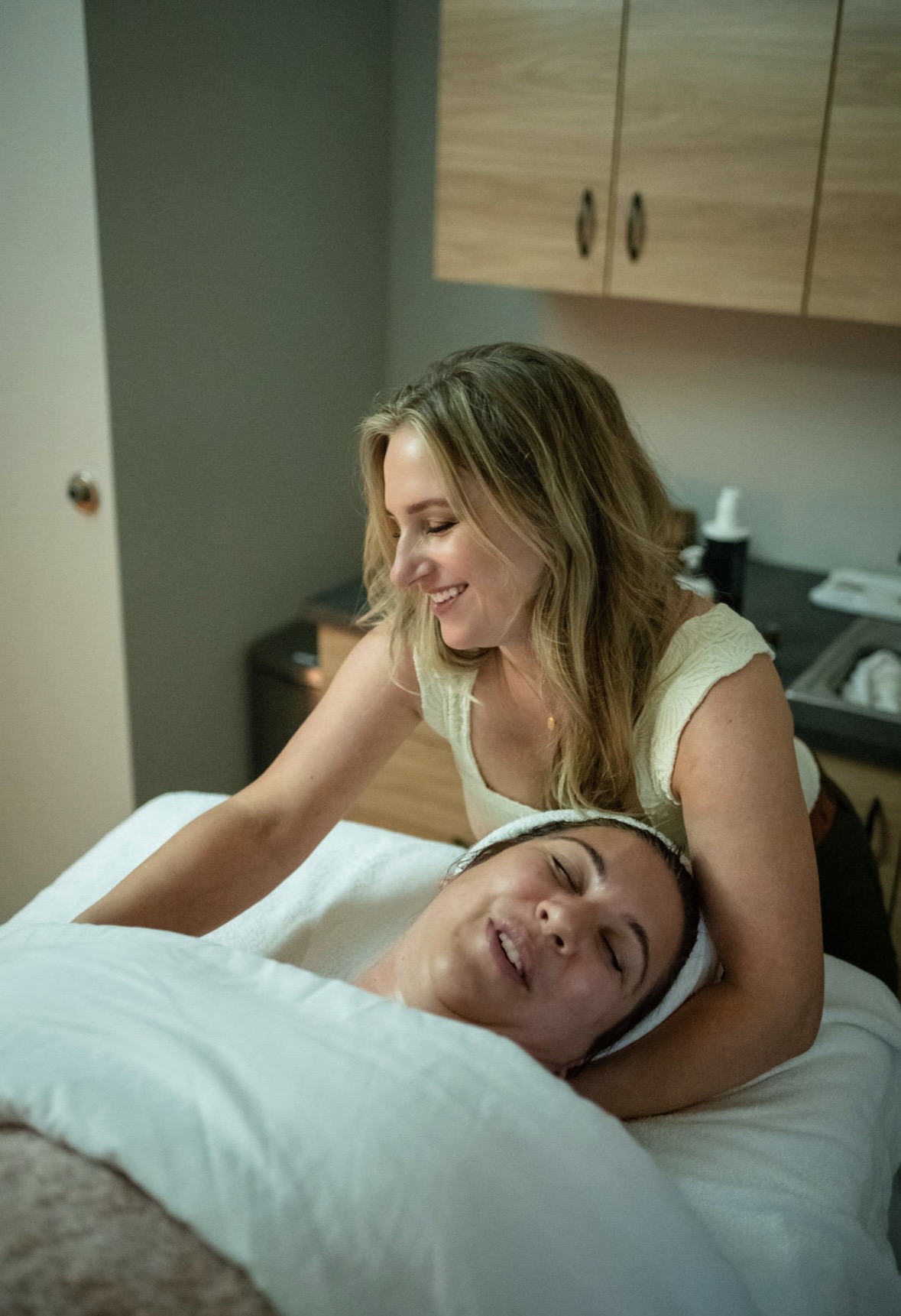 Woman smiling and leaning over a person lying in bed, in a room with wooden cabinets and a desk.