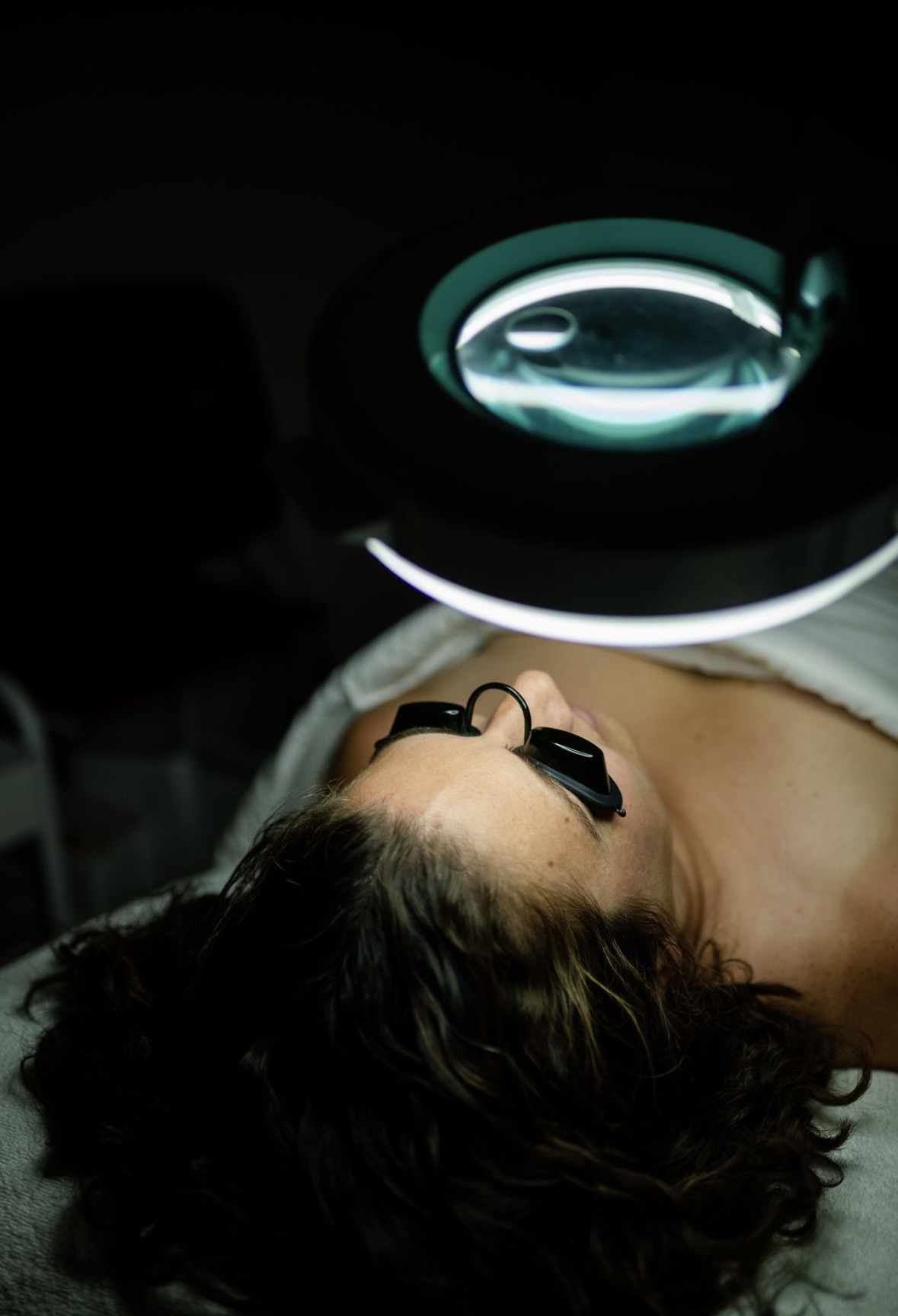 Person lying down with protective eyewear under a circular light, possibly during a medical or dental procedure.