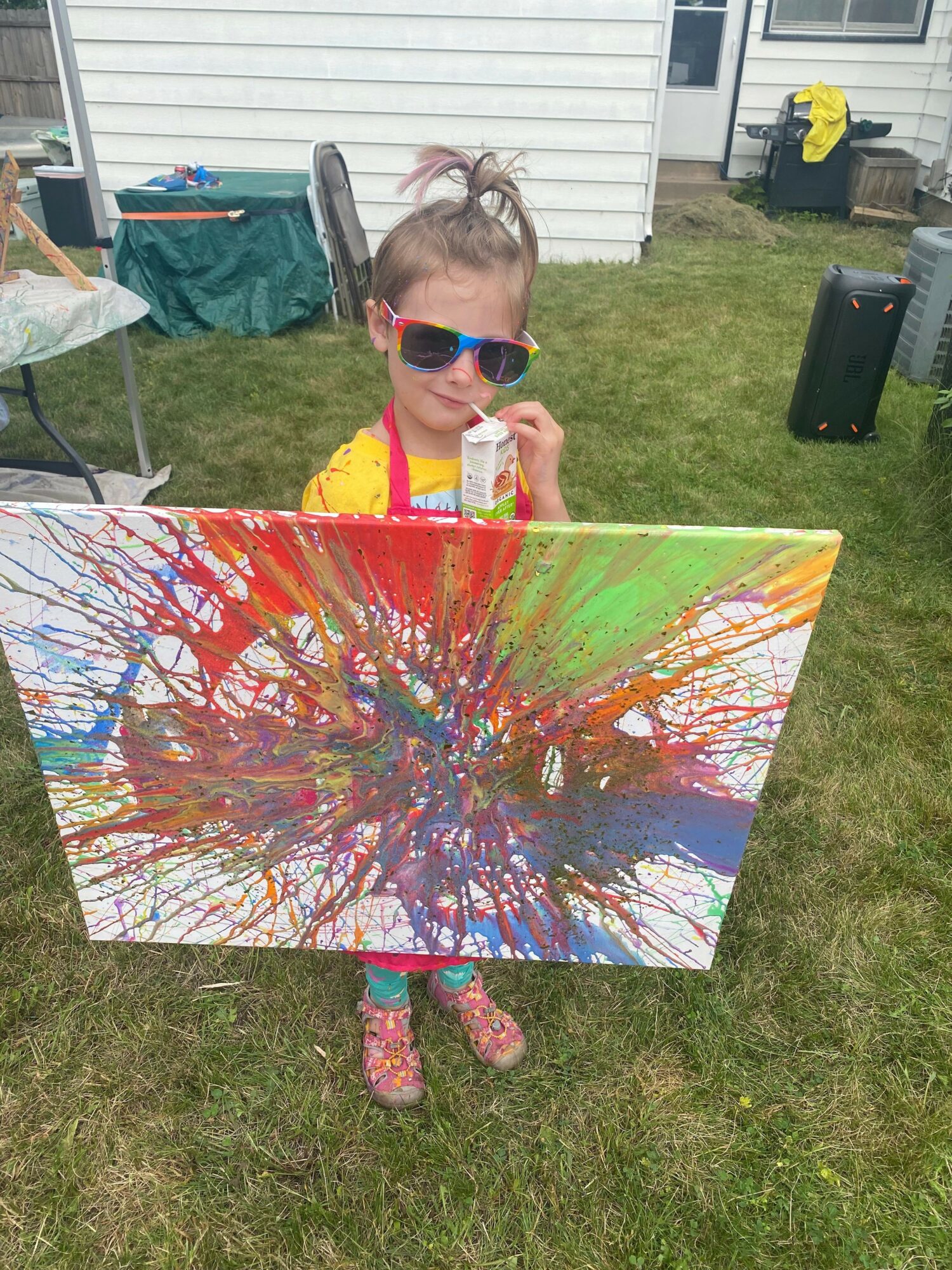 Child wearing sunglasses and yellow shirt stands outdoors next to a colorful splatter painting on a table.