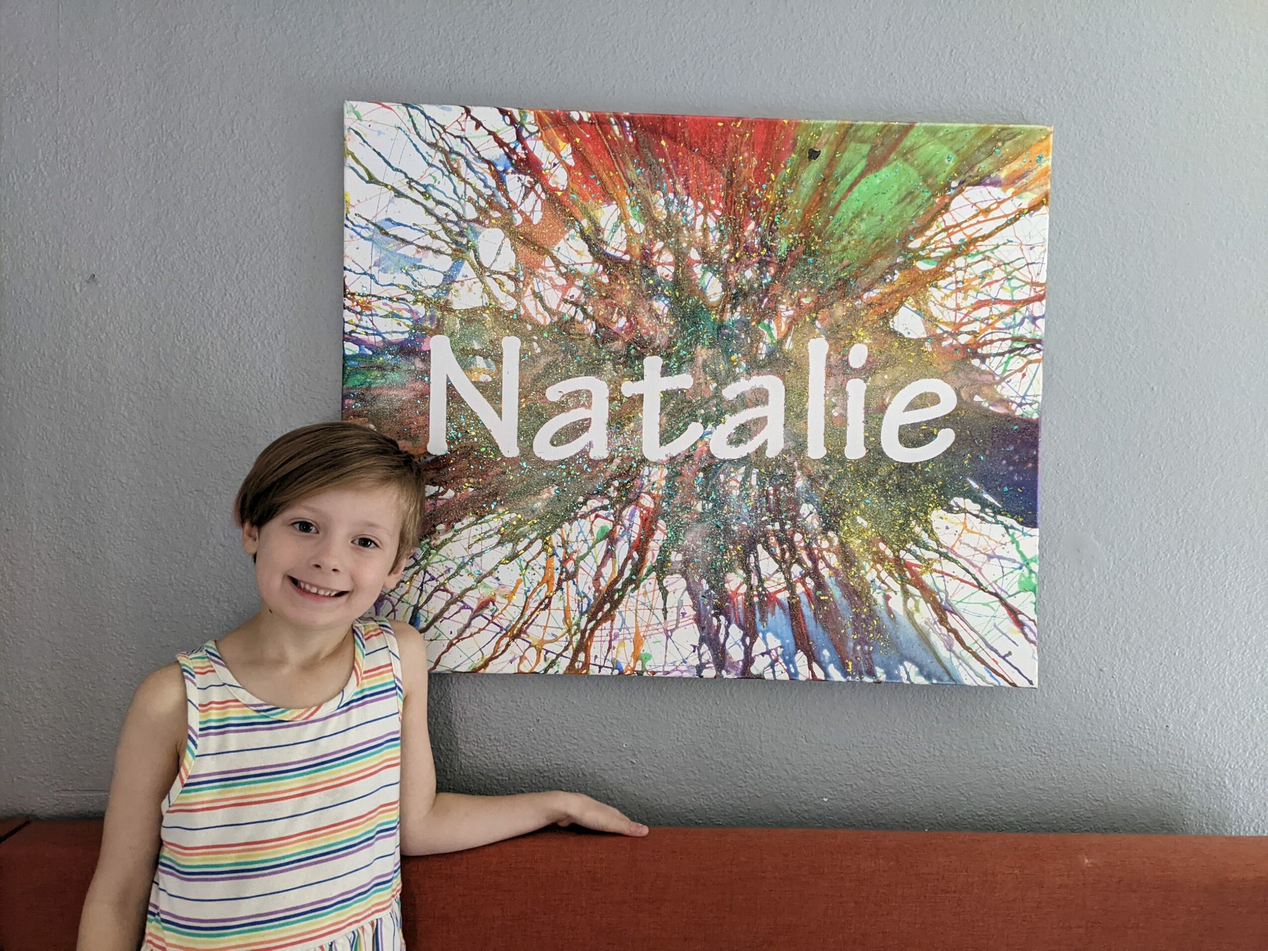 Child with short hair and striped sleeveless shirt standing next to a colorful abstract painting with the name 'Natalie' on it, on a gray wall.