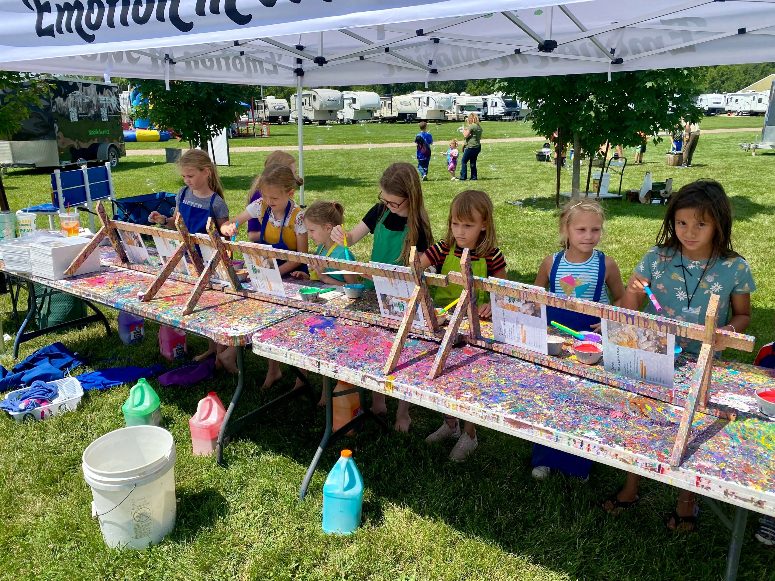 Seven children stand behind a table with colorful artwork, under a white canopy in a park, with trees and people in background.