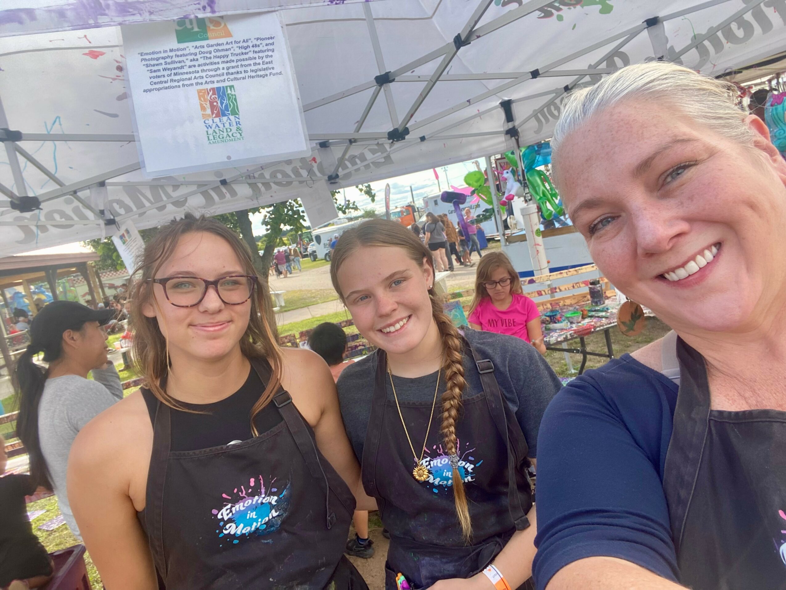 Three smiling people, two young women and an older woman, standing outdoors under a tent, wearing black aprons, at an event.