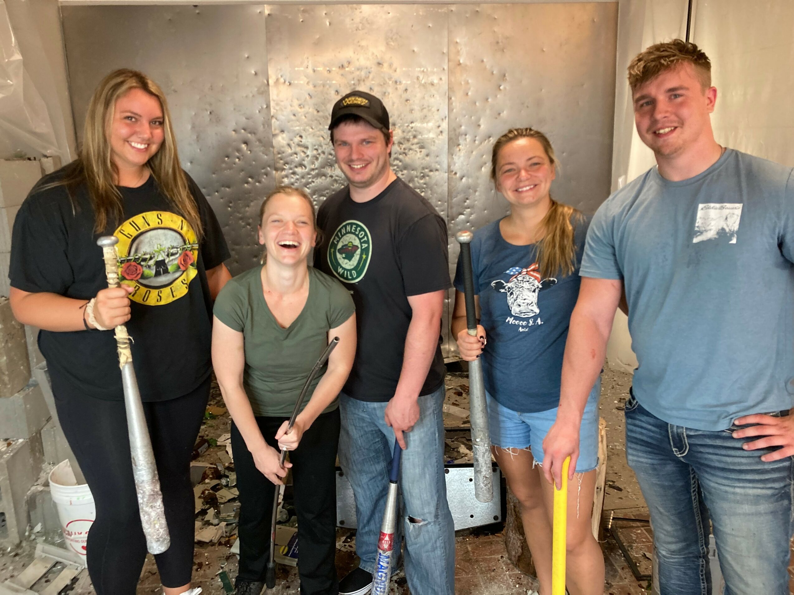 Group of five smiling people standing indoors, some holding yellow and black objects, in front of a textured wall.