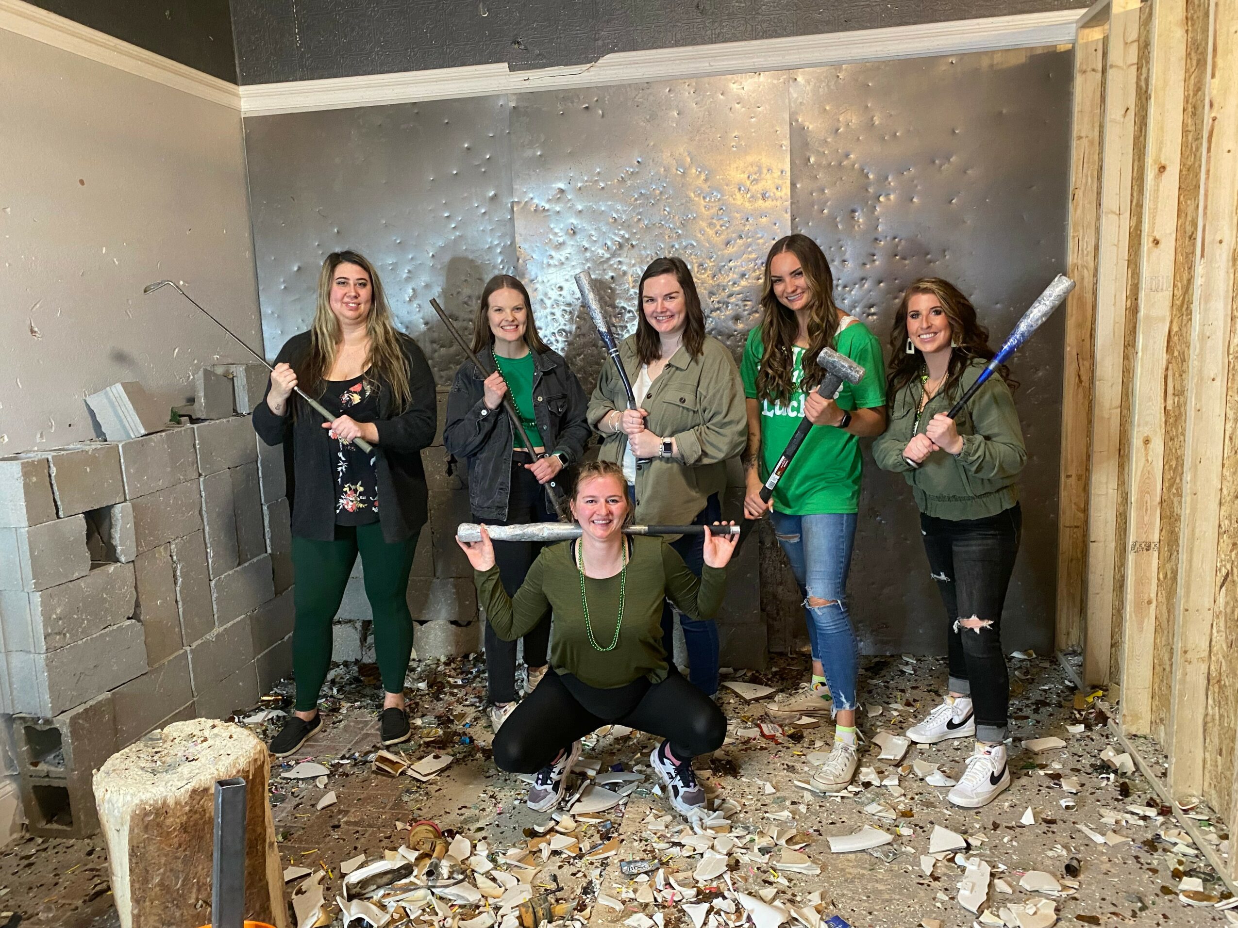Six women in casual clothing pose with sledgehammers in a partially constructed room with debris and cinder blocks.
