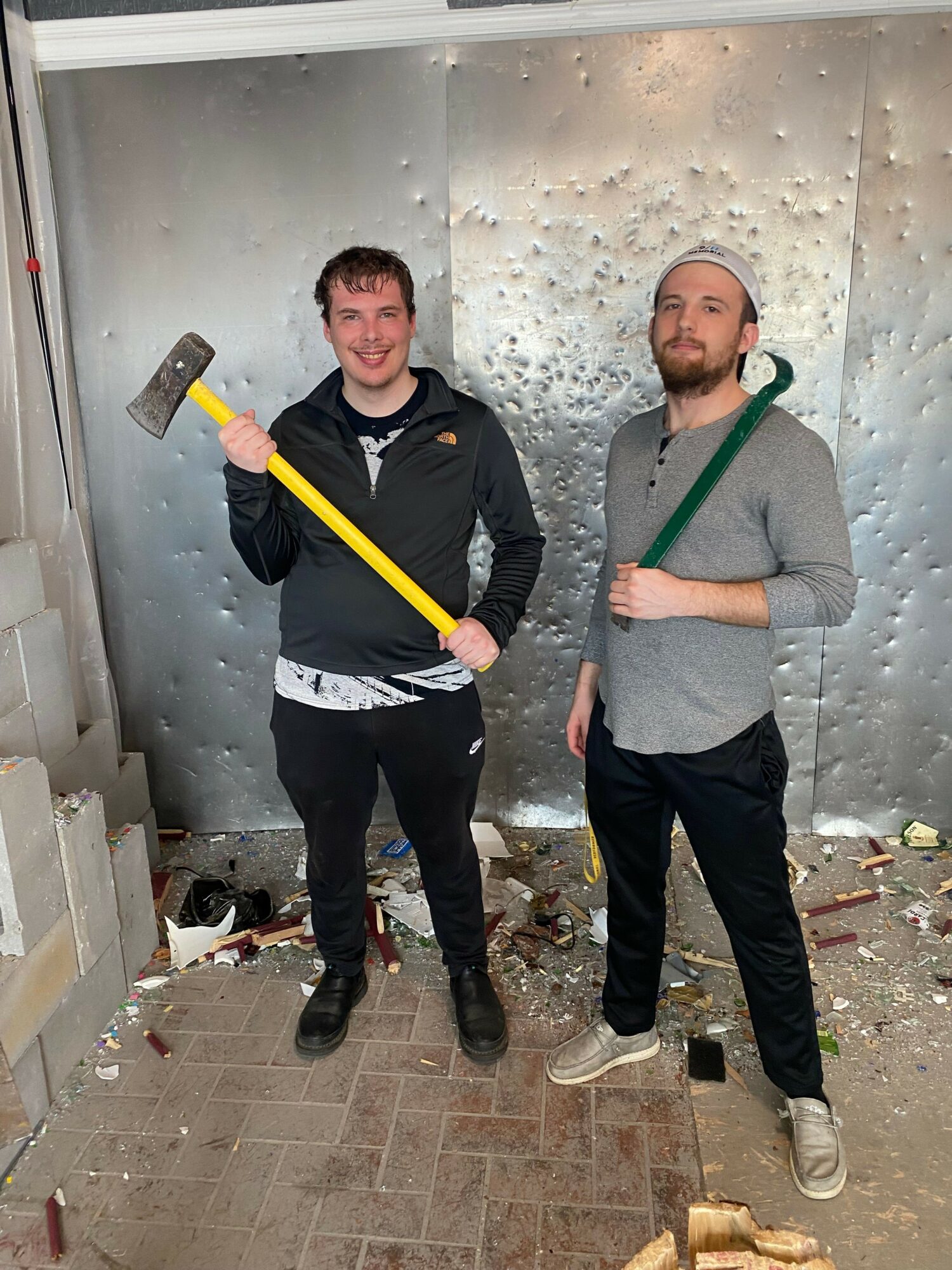 Two men standing indoors, one holding a yellow axe, the other holding a green stick, with debris on the floor.