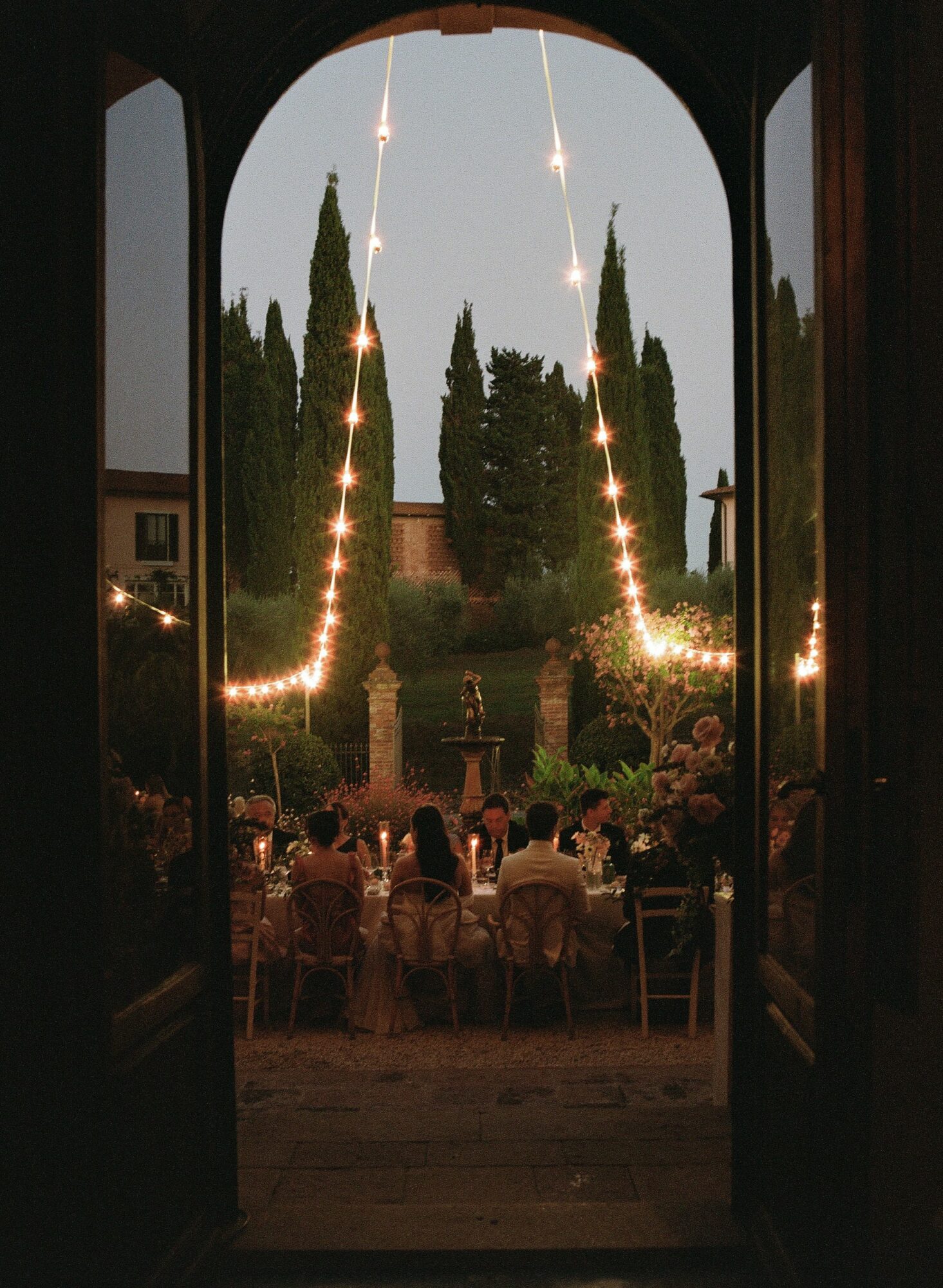 People dining outdoors at sunset, viewed through an arched doorway with trees and string lights in the background.