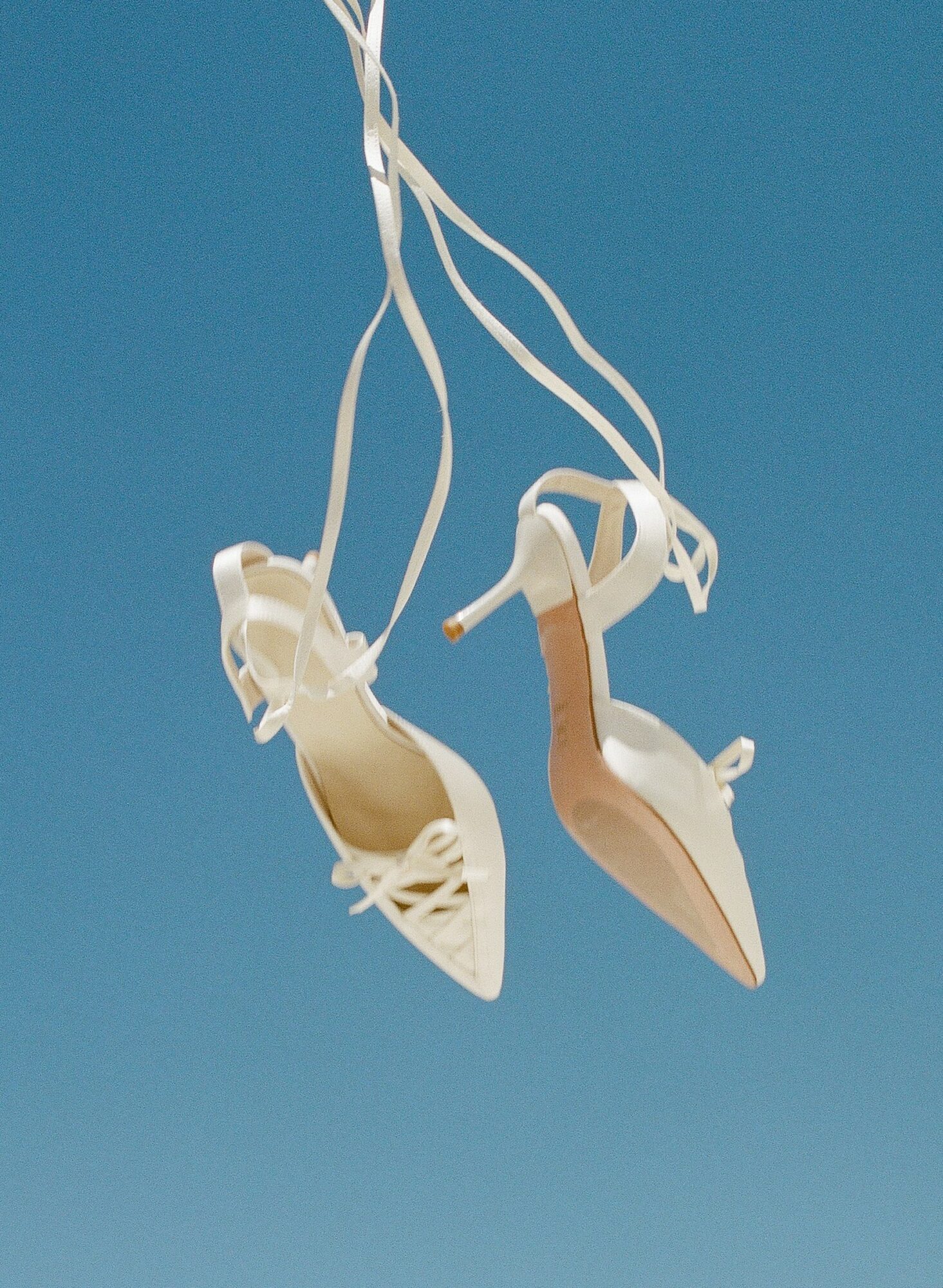 Pair of beige high-heeled shoes hanging by white ribbons against a blue background.