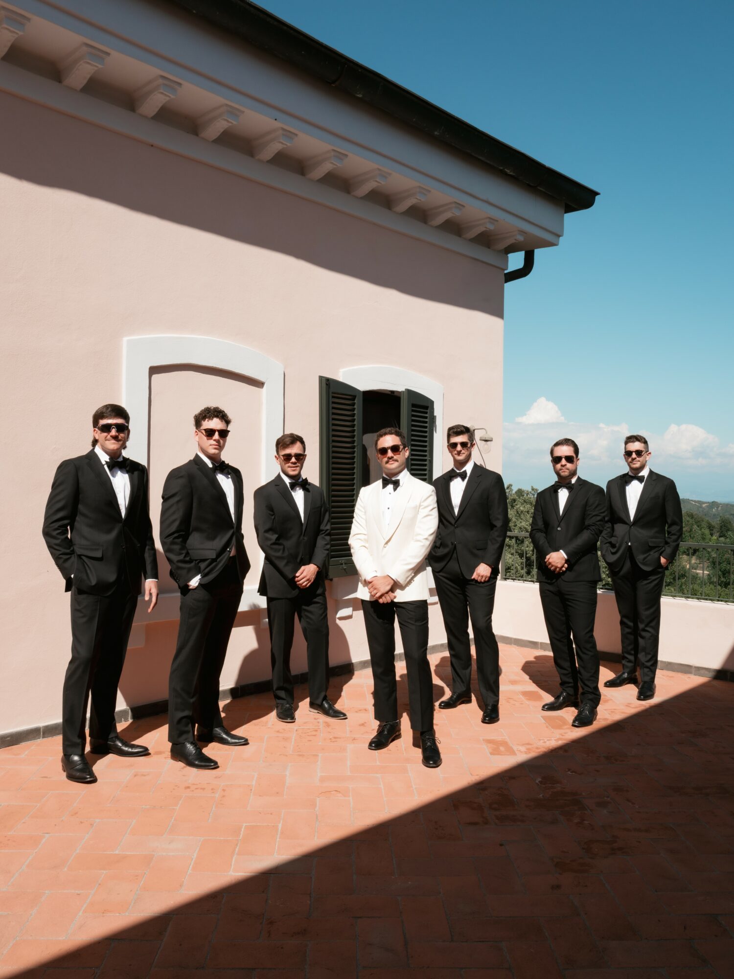 Seven men in tuxedos and one in a white suit stand outdoors on a terrace with a building and blue sky behind them.