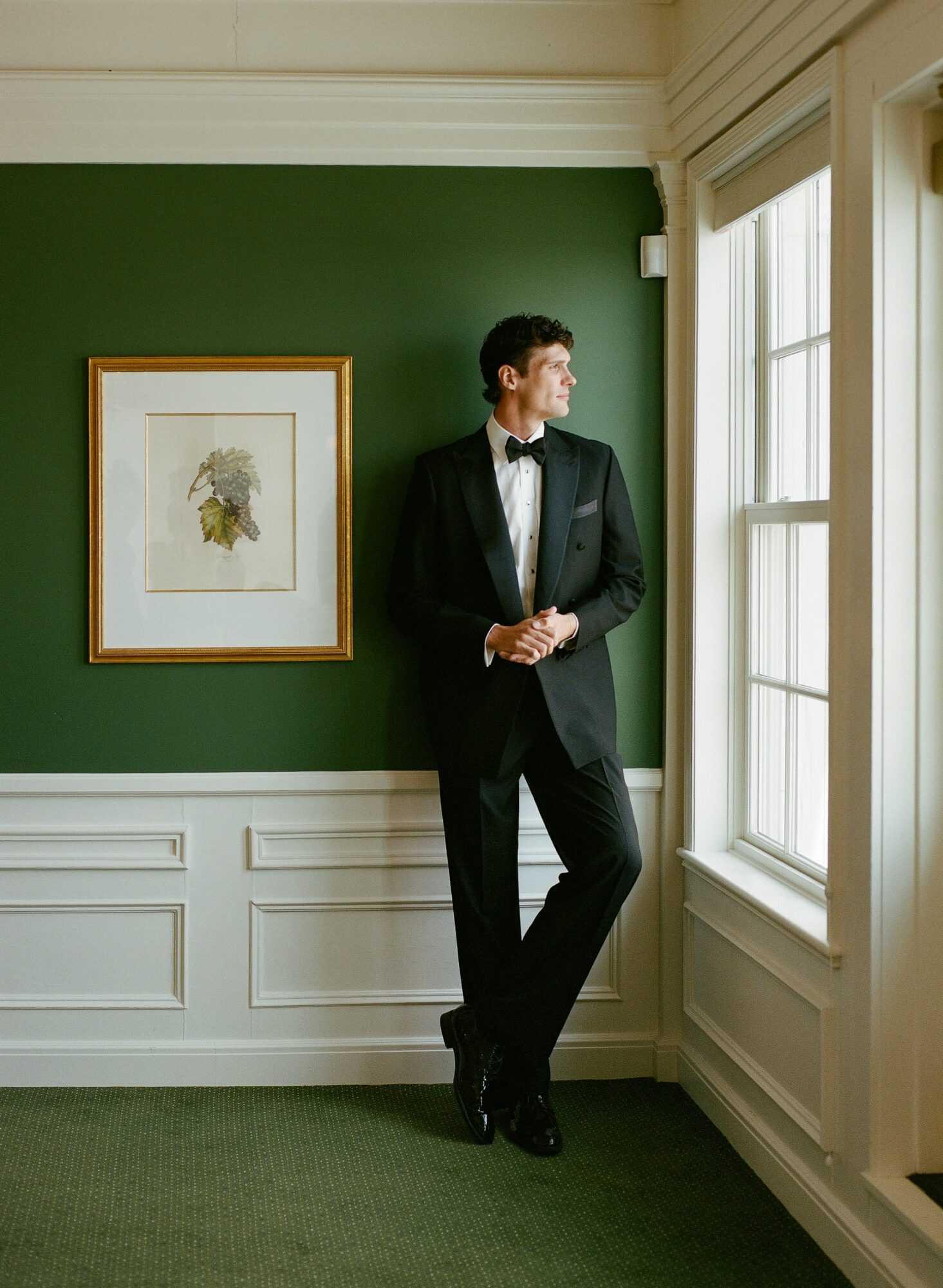 Man in tuxedo standing by window in a room with green wall and framed artwork, looking outside.