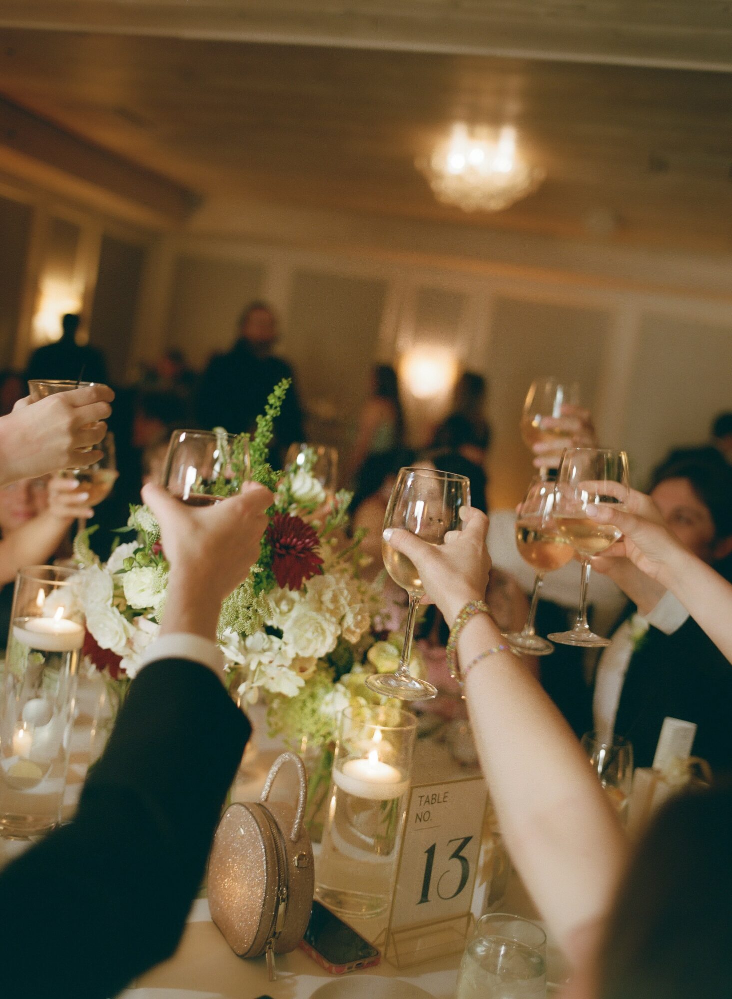 People raising glasses in a toast at a celebration, with floral centerpieces and candles on the table.