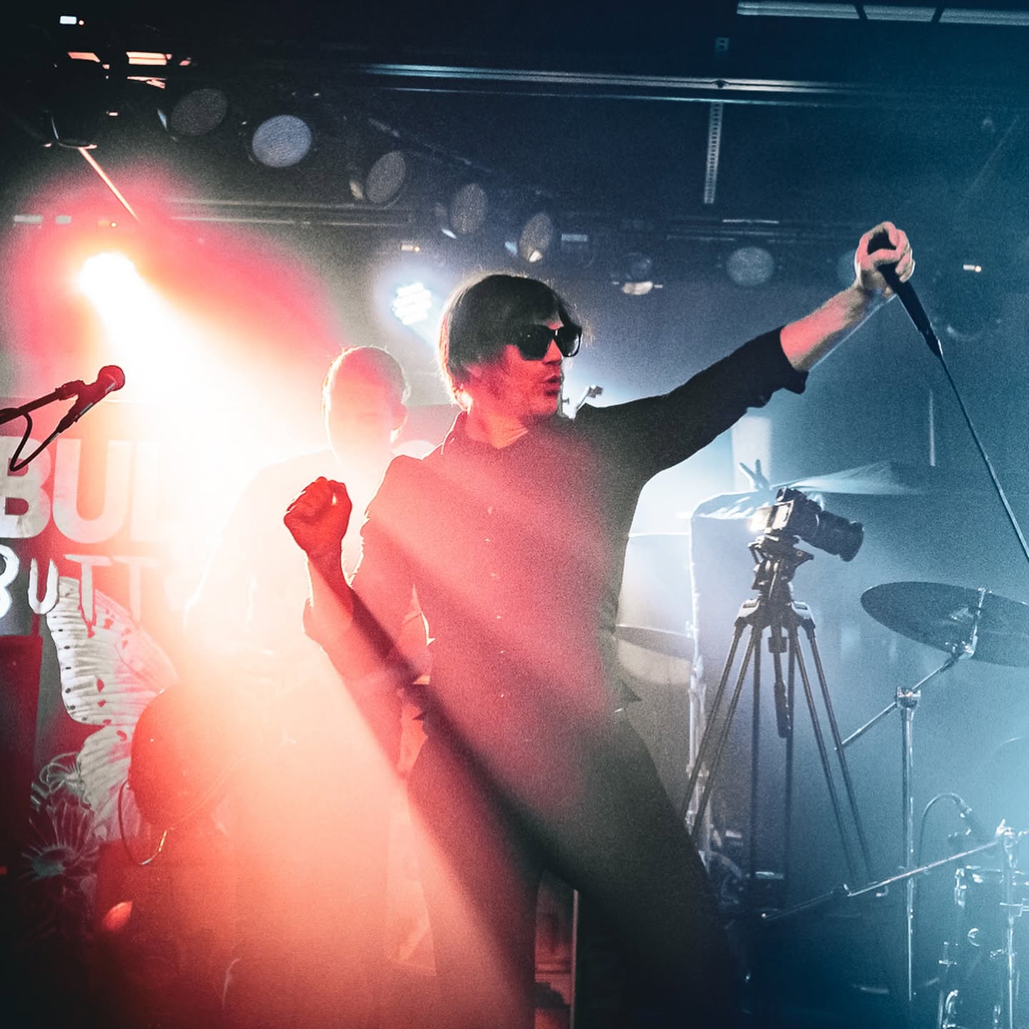 Person with dark hair and sunglasses holding a microphone on stage, with band equipment and colorful stage lights.